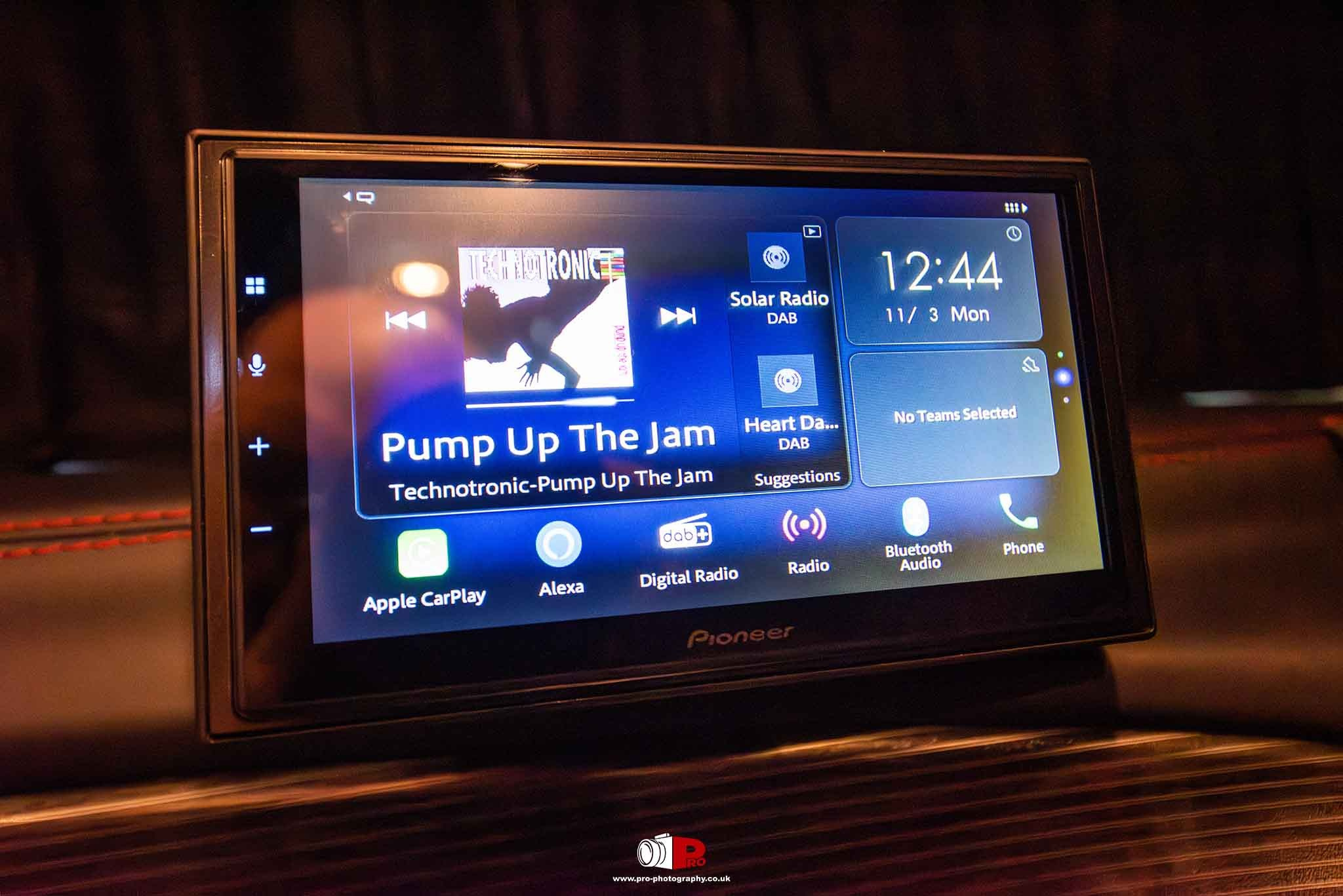 Close-up of a Pioneer car stereo screen displaying "Pump Up The Jam" by Technotronic with various app icons like Apple CarPlay and Alexa.