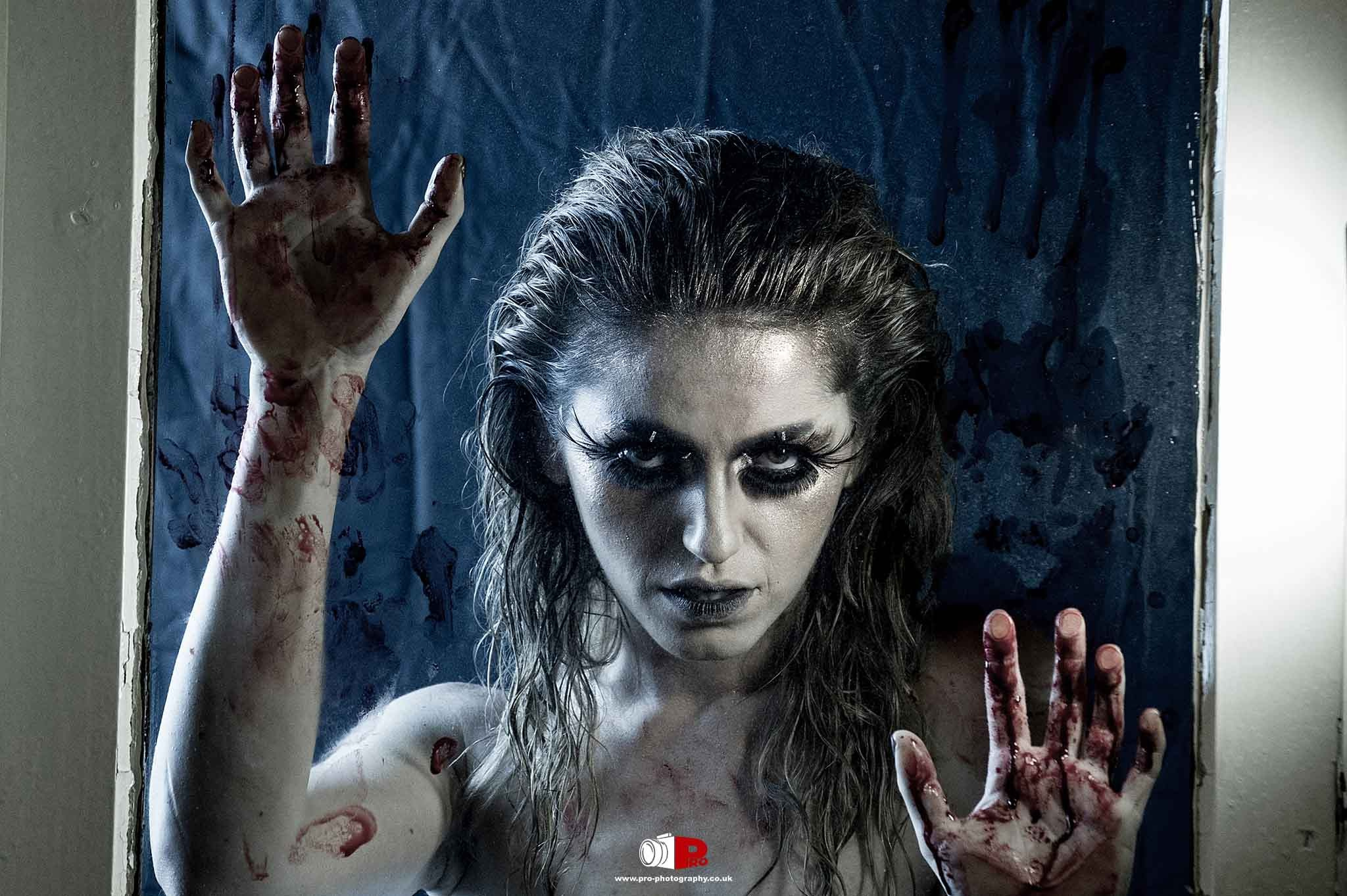 A haunting woman with smeared blood and dark makeup posing against a blood-stained window.