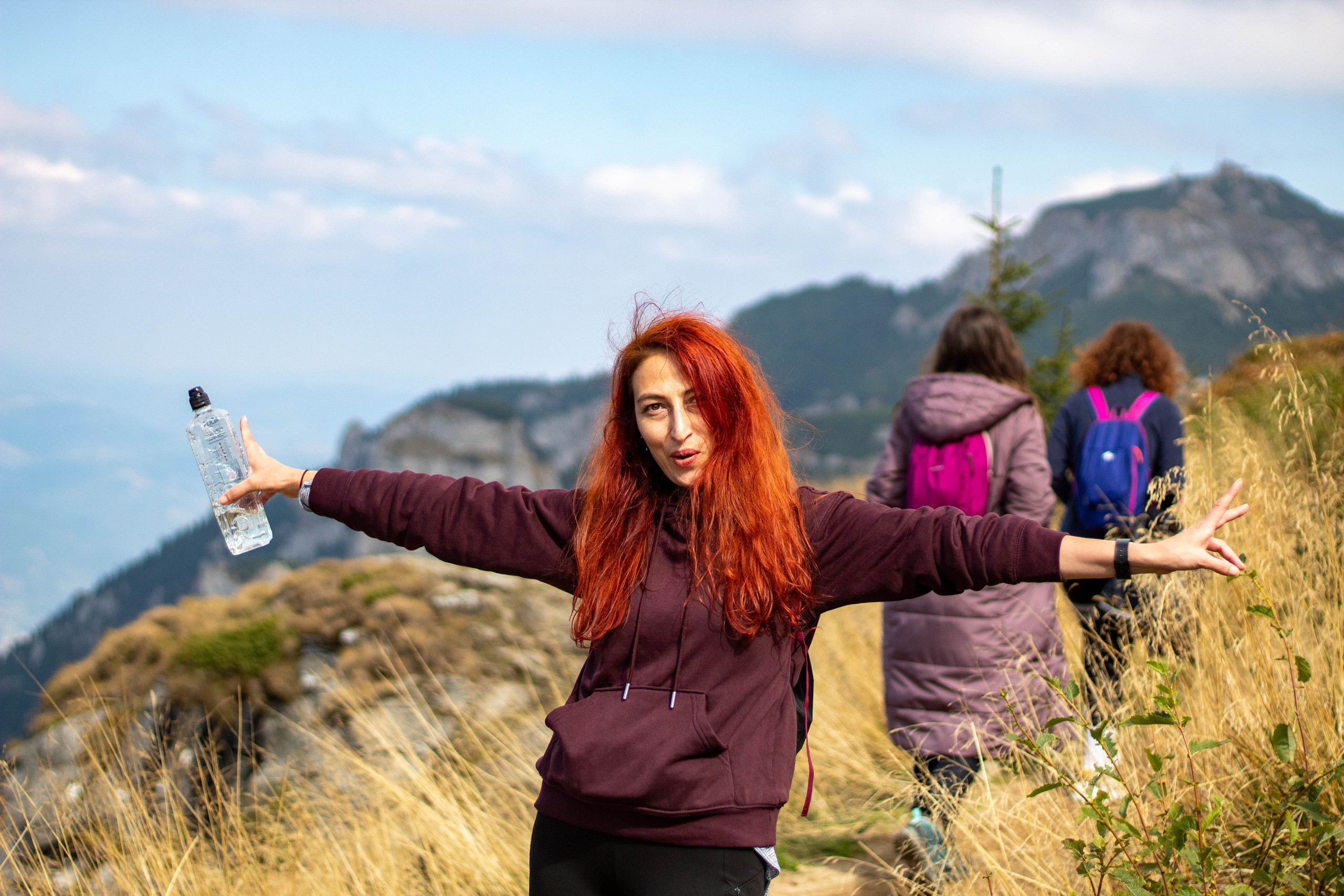 Woman with red hair standing with open arms on a mountain trail, scenic peaks and sky in the background.