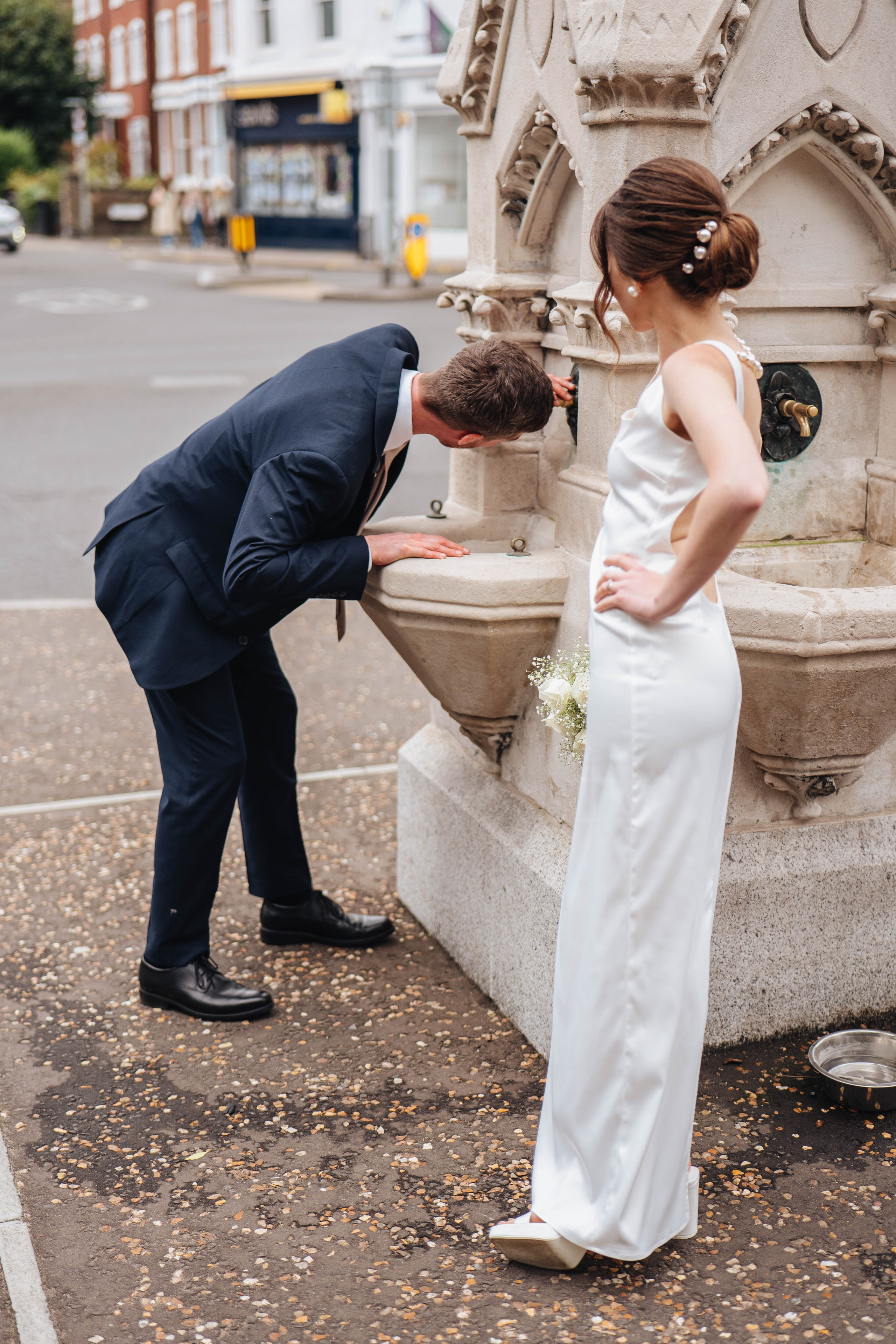 groom drinking water from fountain while bride is waiting