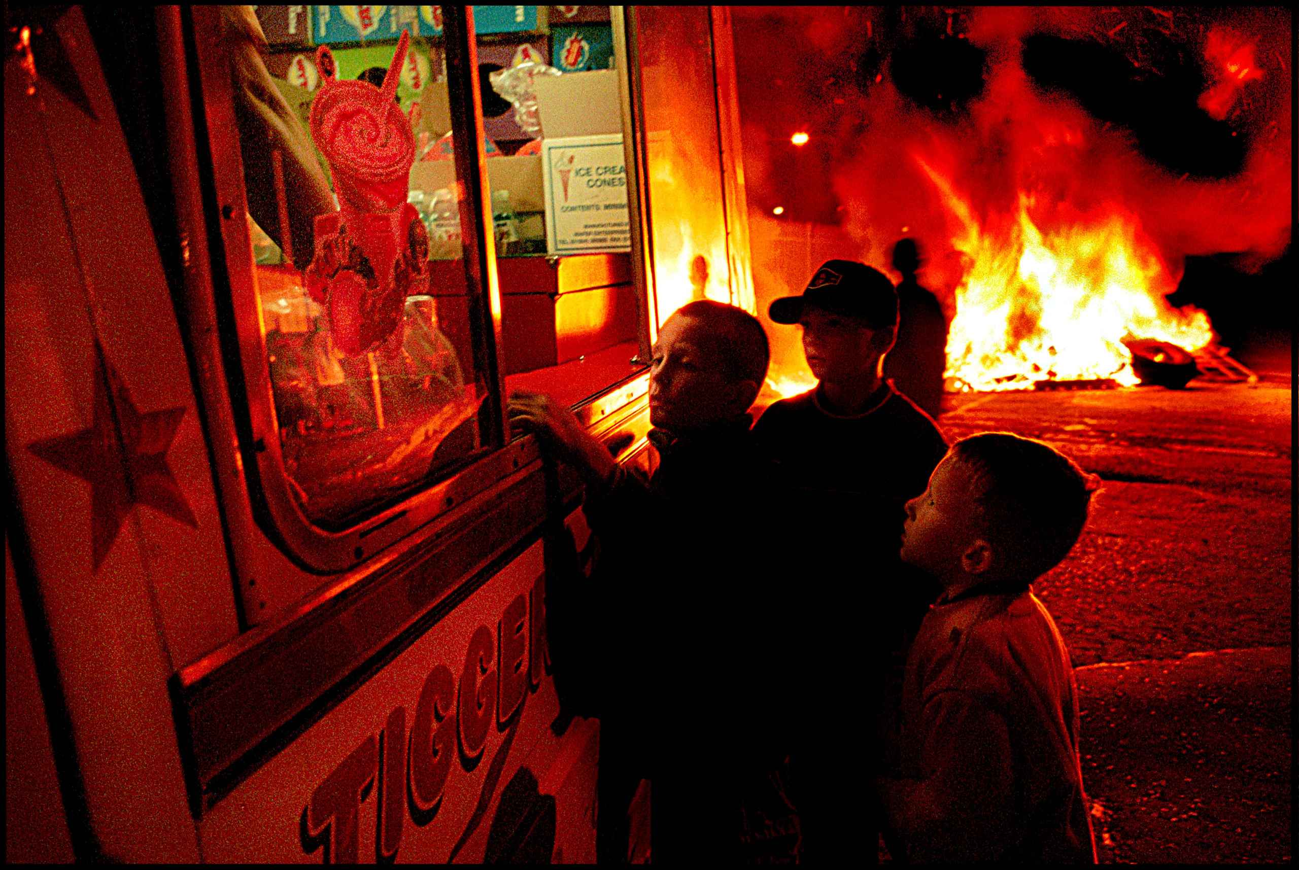 Children in Belfast's Sandy Hill neighborhood queue for ice cream as a road block burns in the background.