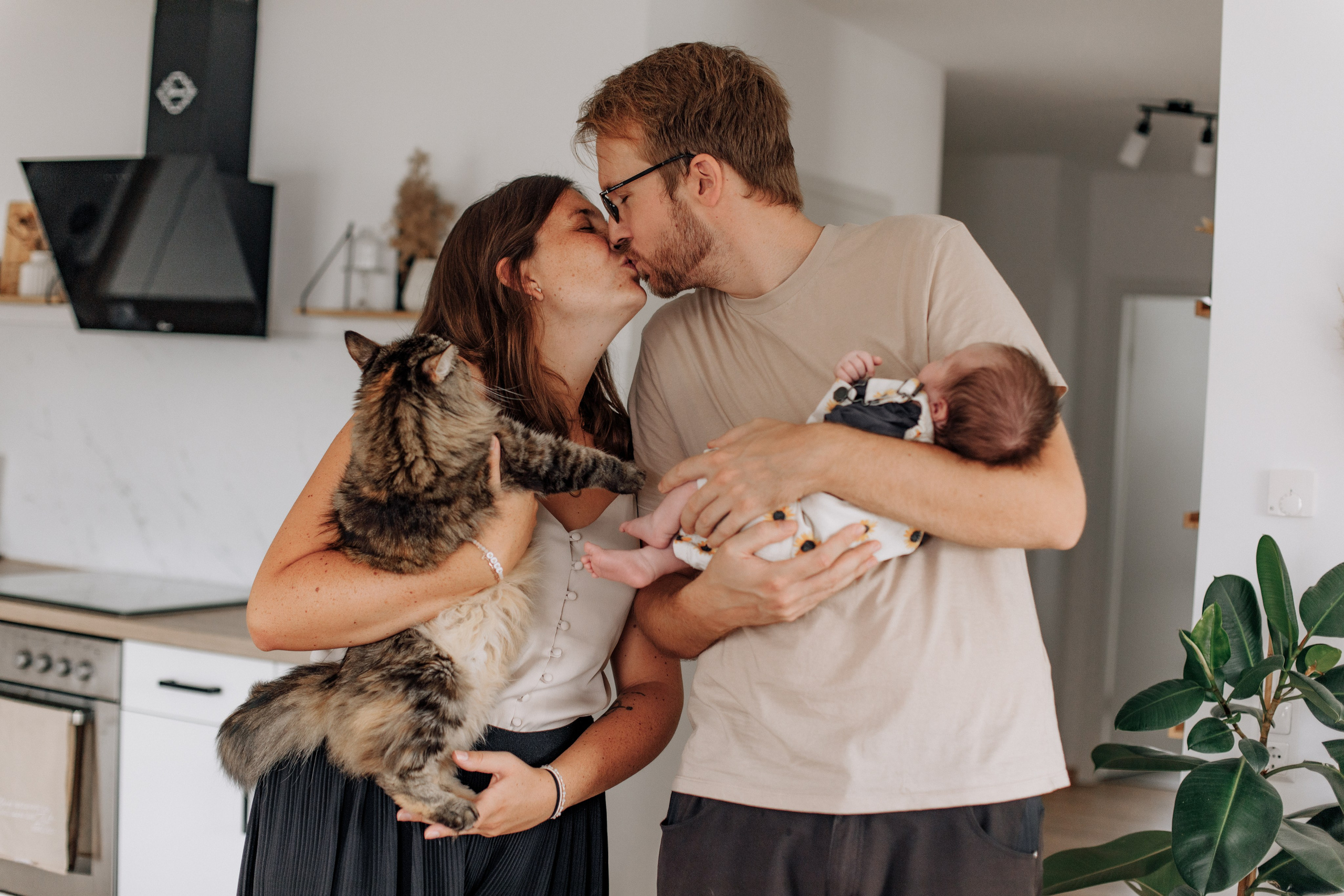 Familienporträt: Mutter hält Katze, Vater hält Baby, beide küssen sich liebevoll.