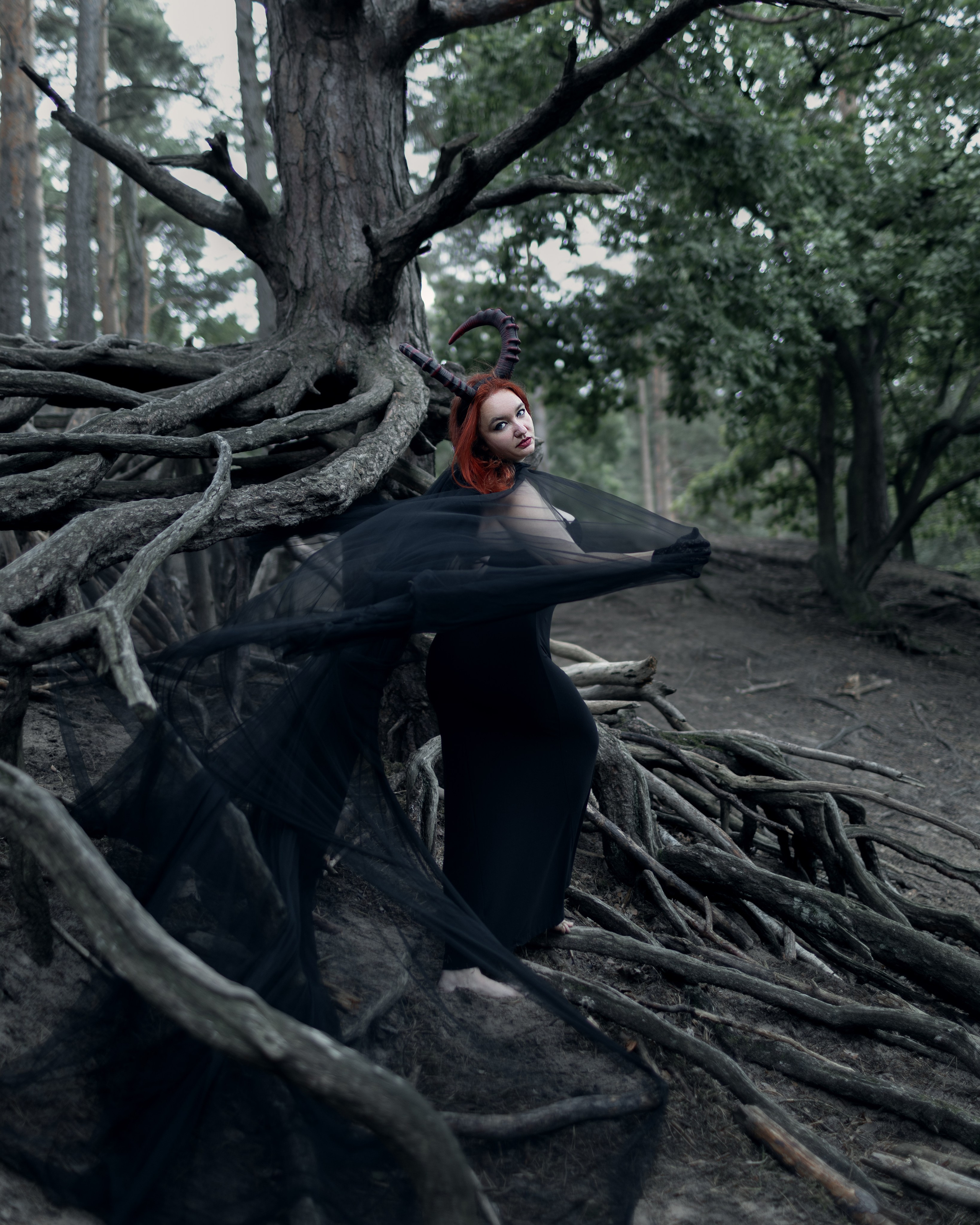 Ksenia steht mit Teufelshörnern und schwarzem Umhang auf Baumwurzeln im düsteren Wald