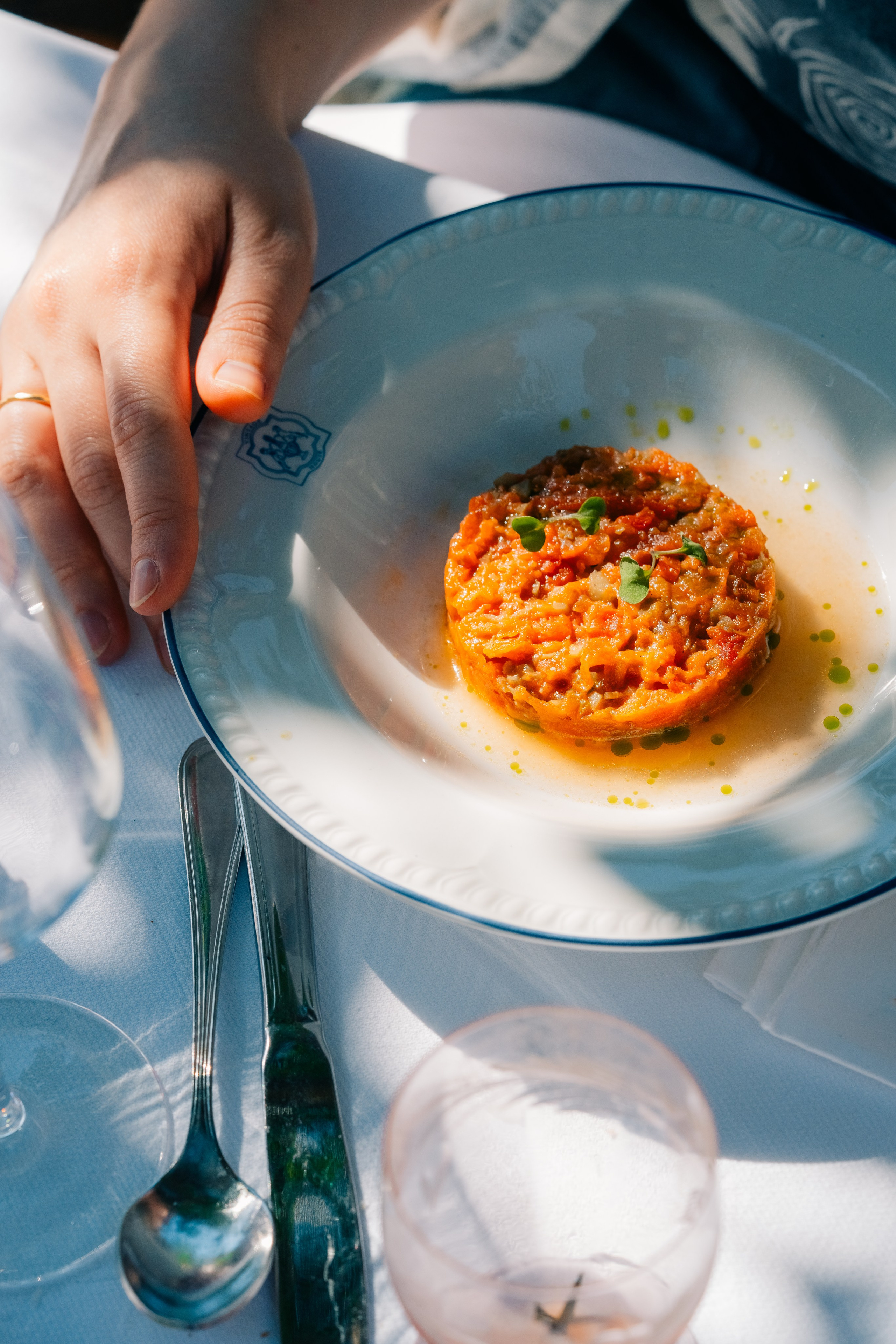 Mão a alcançar um prato de prato gourmet servido ao ar livre. Mão a alcançar um prato gourmet de legumes em cubos finos servidos num prato branco, ao ar livre numa mesa iluminada pelo sol.