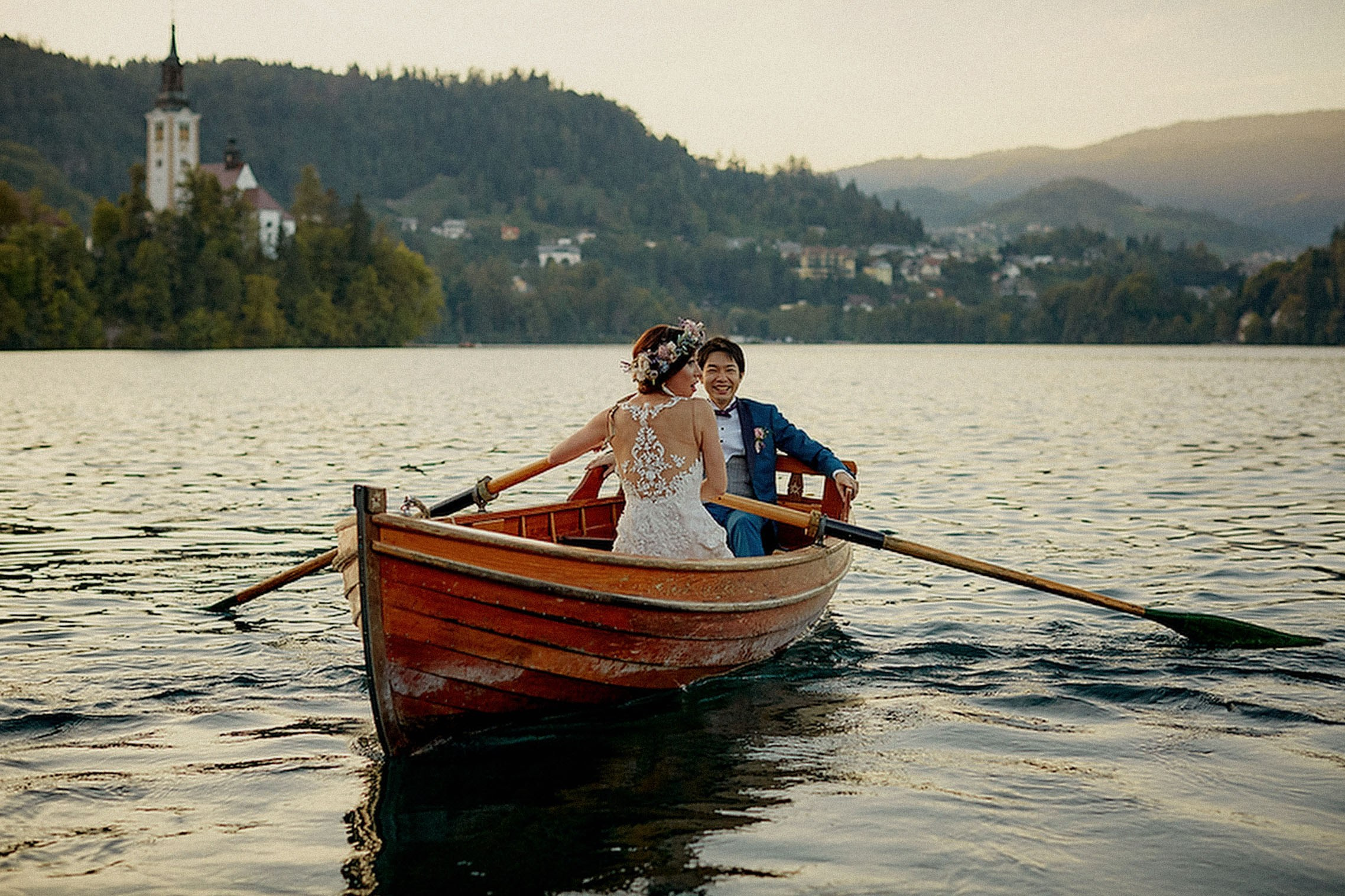 A Japanese bride wearing a floral headpiece sits in a boat as her groom rows using its oars.