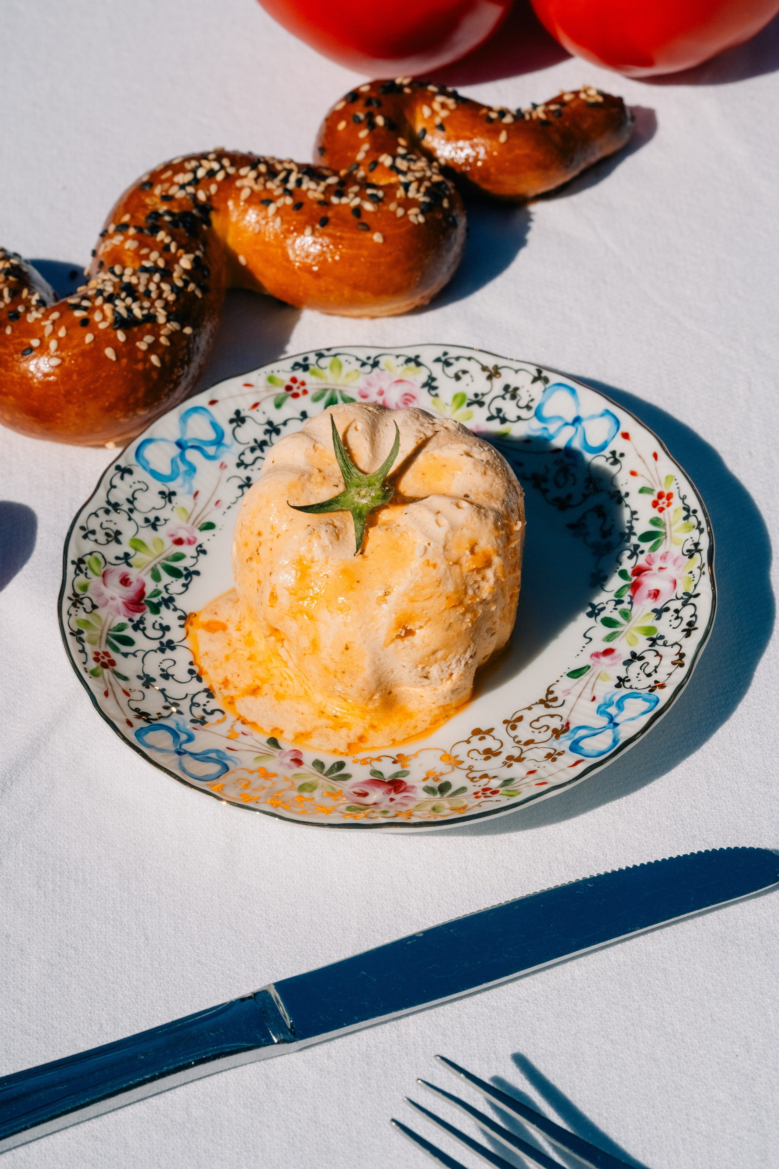 Arranjo alimentar artístico de tomate em prato decorativo com pão. Arranjo alimentar exclusivo com um tomate num prato floral decorativo, acompanhado por um pretzel coberto de sésamo, colocado sobre uma mesa iluminada pelo sol.