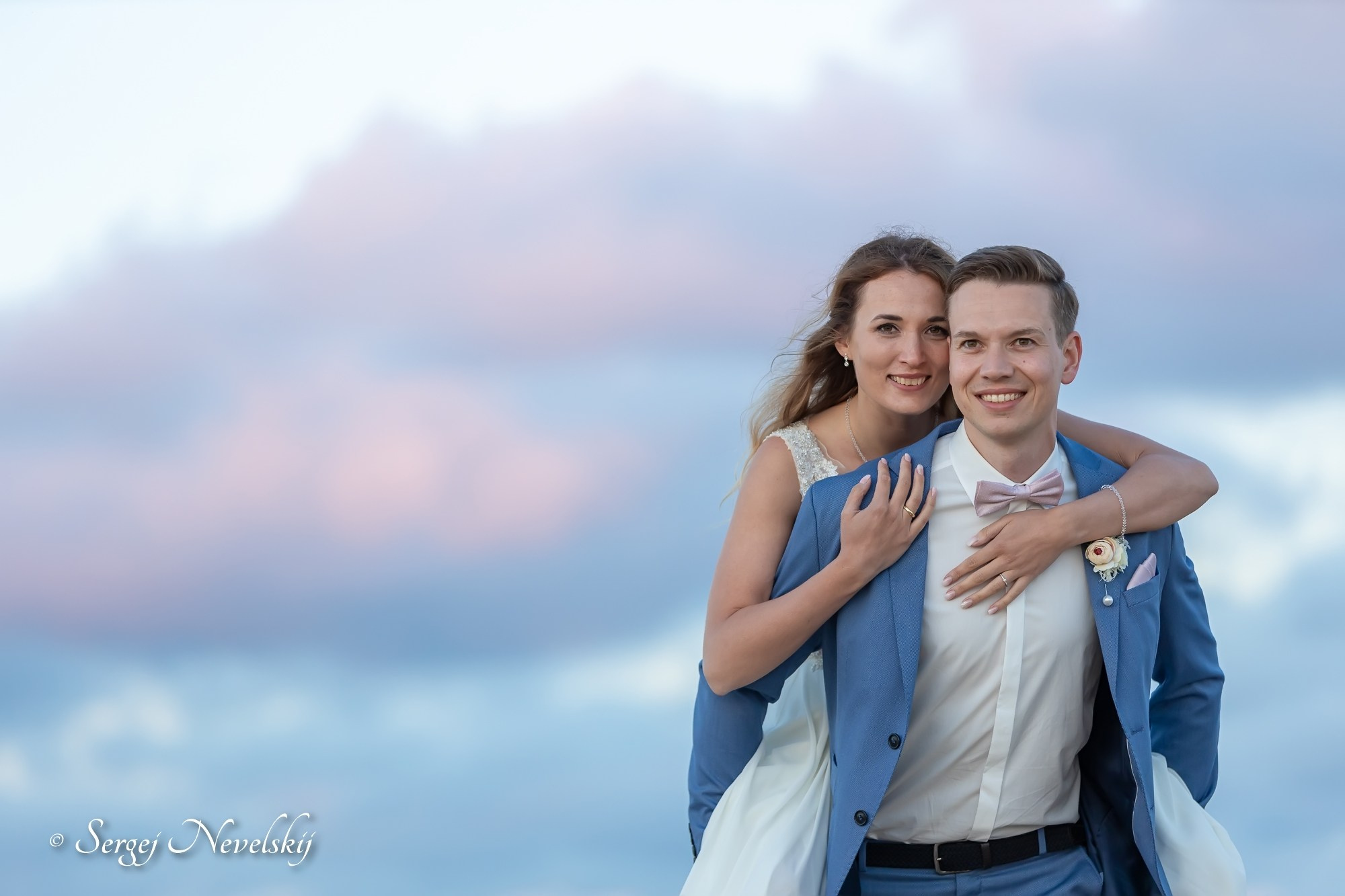 English:Joyful newlyweds embracing against a dreamy pastel sky at golden hour. Bride in a delicate lace-top white wedding dress, groom in a light blue suit with matching jacket draped over her shoulders, pink bow tie and rose boutonniere. Both smiling radiantly, capturing pure happiness and love. Photo by © Sergej NevelskijРусский:Счастливые молодожёны обнимаются на фоне нежного пастельного неба в час заката. Невеста в белом свадебном платье с кружевным верхом, жених в голубом костюме, накинувший пиджак ей на плечи, с розовой бабочкой и бутоньеркой. Оба сияют от счастья — чистая любовь и радость. Фото © Sergej NevelskijDeutsch:Glückliches Brautpaar umarmt sich vor einem traumhaften pastellfarbenen Himmel in der Golden Hour. Braut im weißen Brautkleid mit Spitzenoberteil, Bräutigam im hellblauen Anzug, dessen Jackett er ihr über die Schultern gelegt hat, mit rosa Fliege und Rosen-Anstecker. Beide strahlen vor Freude – pure Liebe und Glück. Foto © Sergej Nevelskij