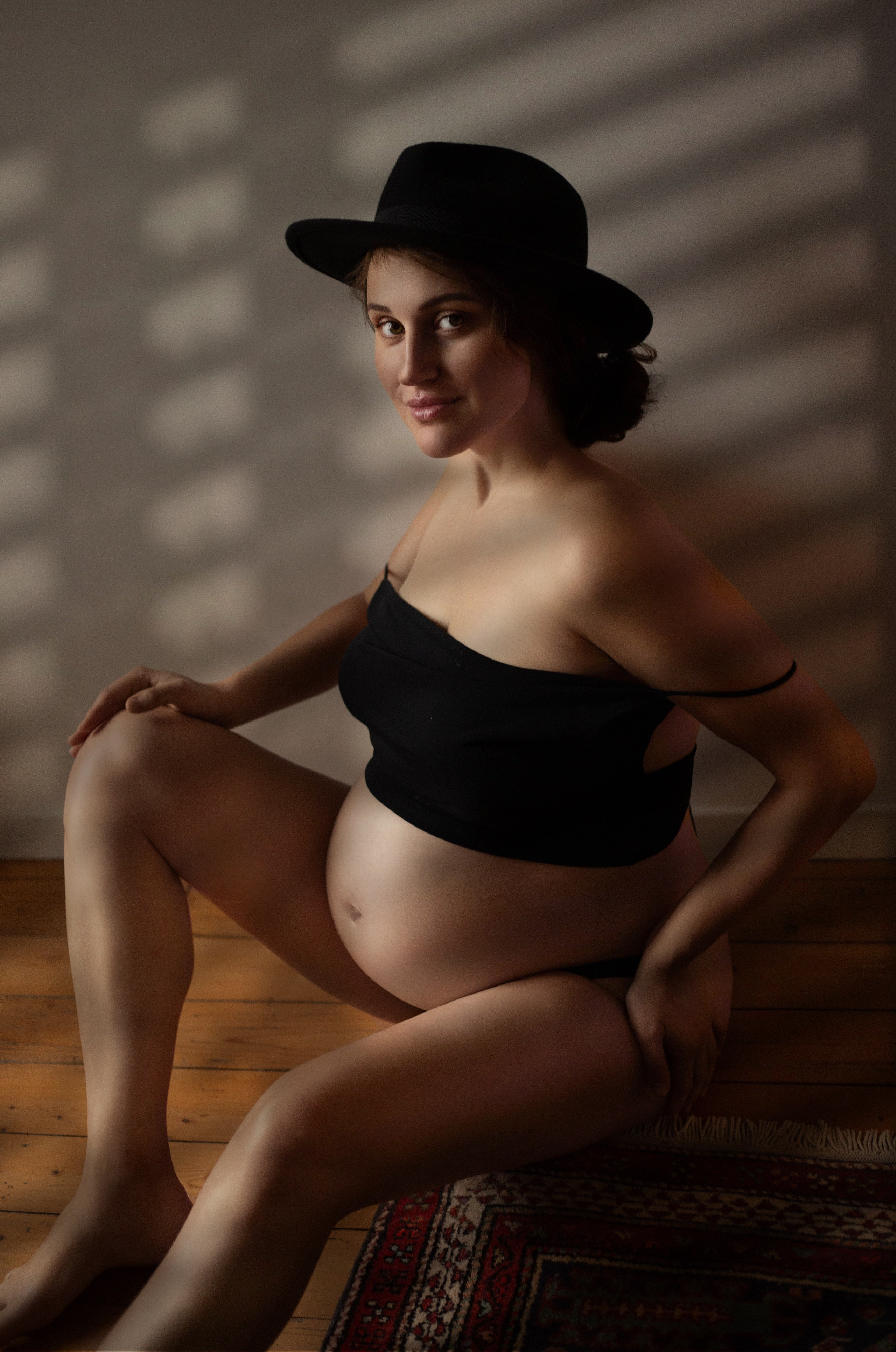 Femme enceinte élégante en tenue noire et chapeau, lumière artistique à travers les stores
