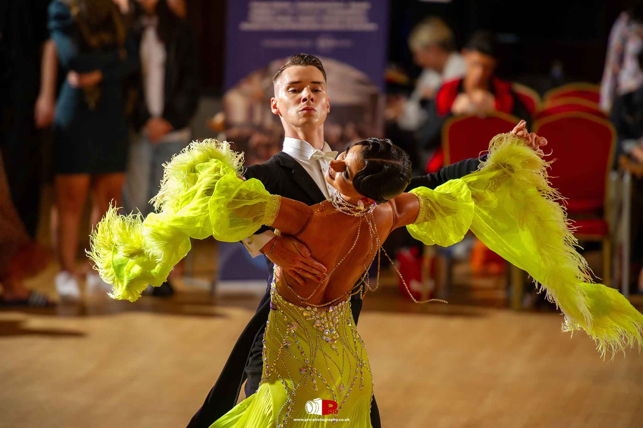 A striking ballroom dance couple performing in a bright yellow feathered costume during a competition.