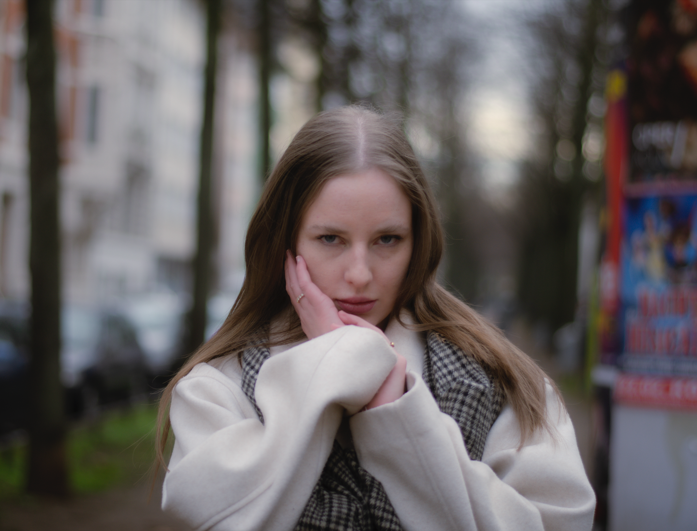 Porträt einer jungen Frau mit dunkelblonden Haaren, blauen Augen und hellbeigem Mantel stehend im Park