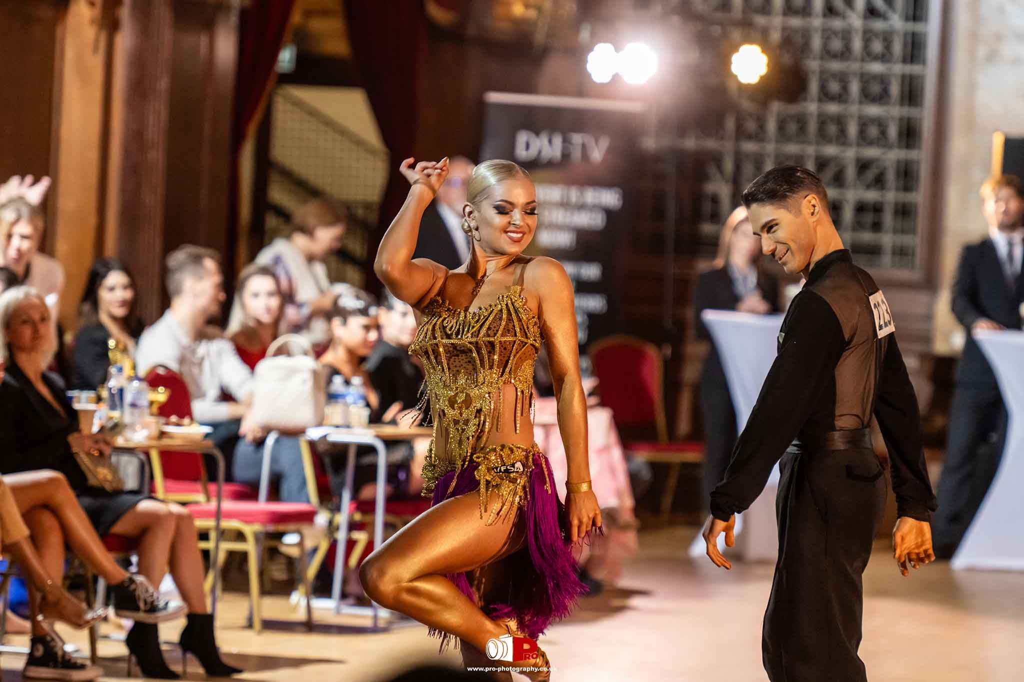 Latin dancers perform a lively routine, with the female dancer in a gold fringe outfit smiling confidently on stage.