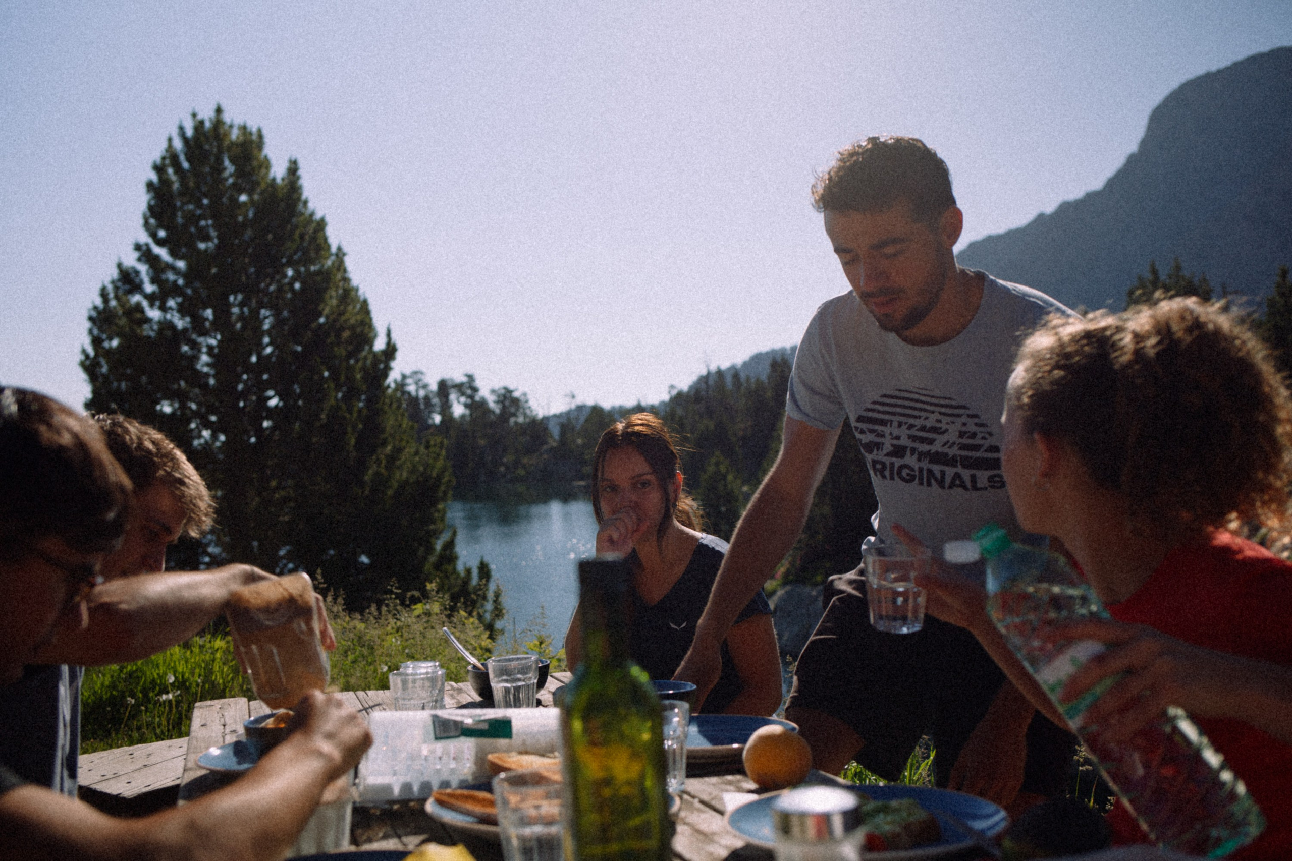 Workers having breakfast outdoors at Josep M. Blanc Refuge with mountains and lake in the background, capturing a natural, unposed moment filled with quiet morning atmosphere and subtle harmony