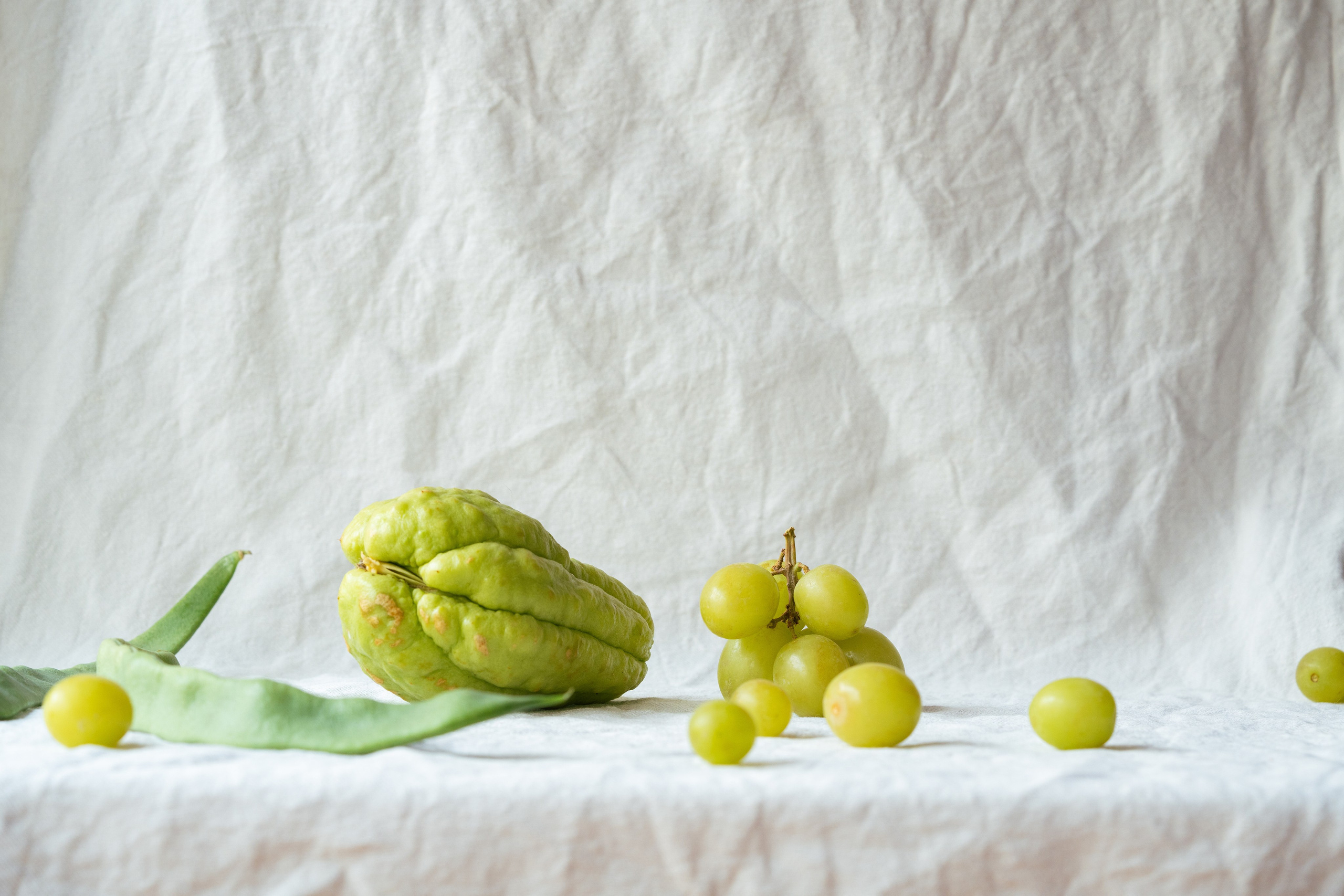 Arranjo de produtos frescos com Chayote e uvas em tecido branco. Composição minimalista de chayote verde fresco, uvas e feijão em um pano de fundo branco texturizado.