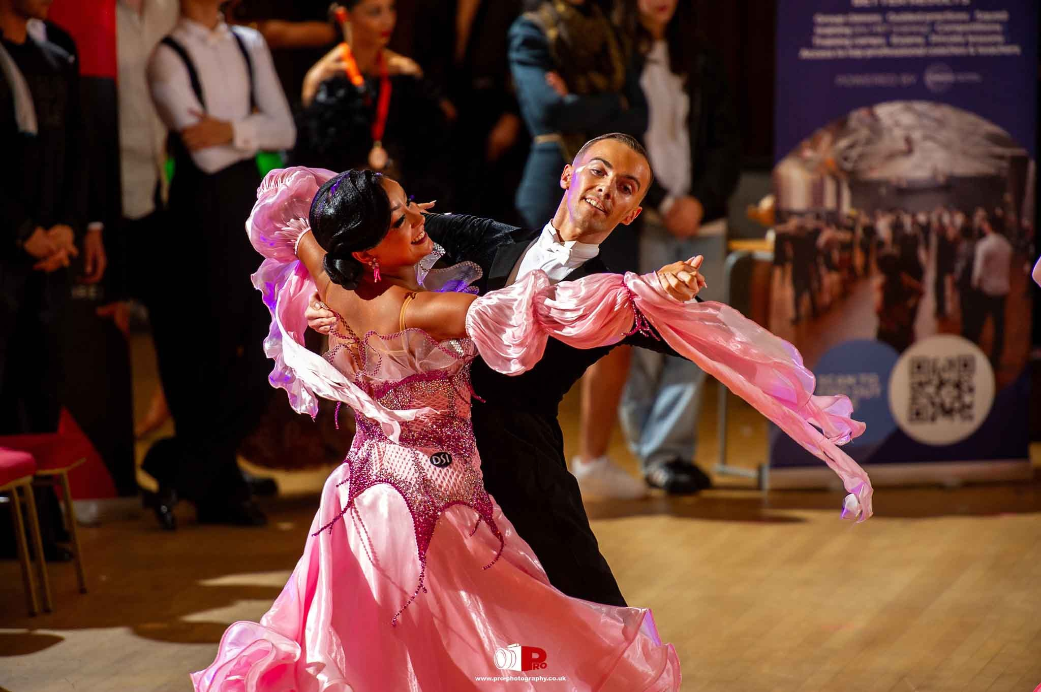 A graceful ballroom dance couple in pink attire performing a captivating dance routine at a competition, surrounded by spectators.