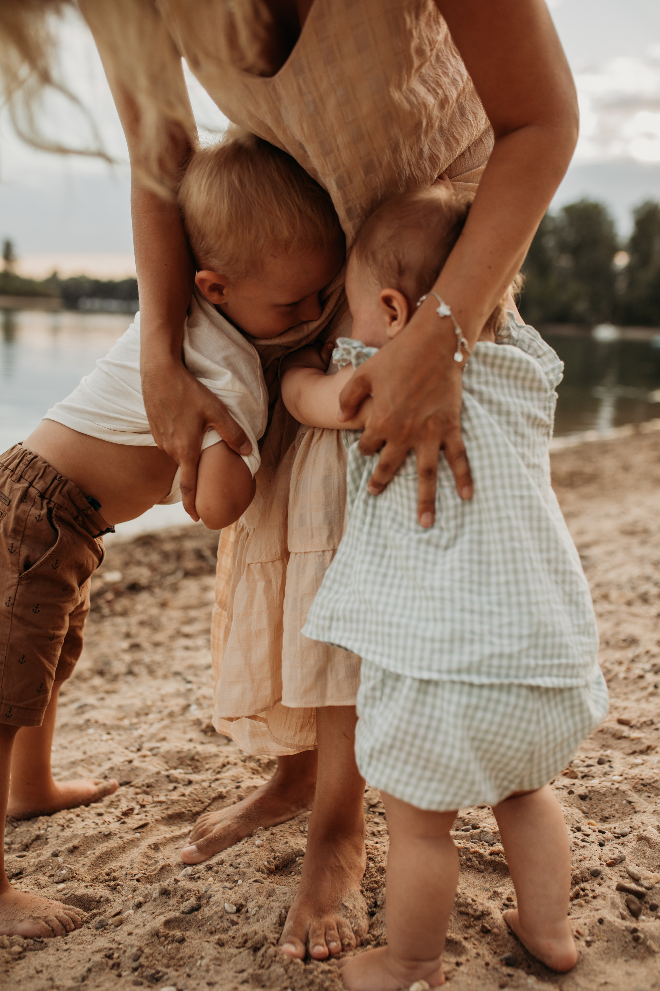 Zwei kleine Kinder umarmen Beine ihrer Mutter  auf dem Strand beim Familienshooting in der Nähe von Mannheim.