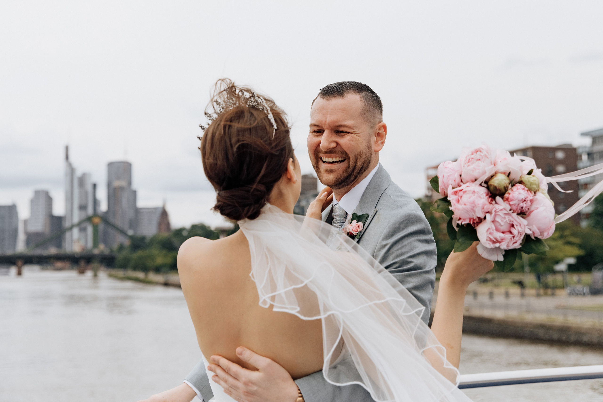 Der Bräutigam umarmt die Braut und lacht mit Blick auf Frankfurt und den Fluss Main.