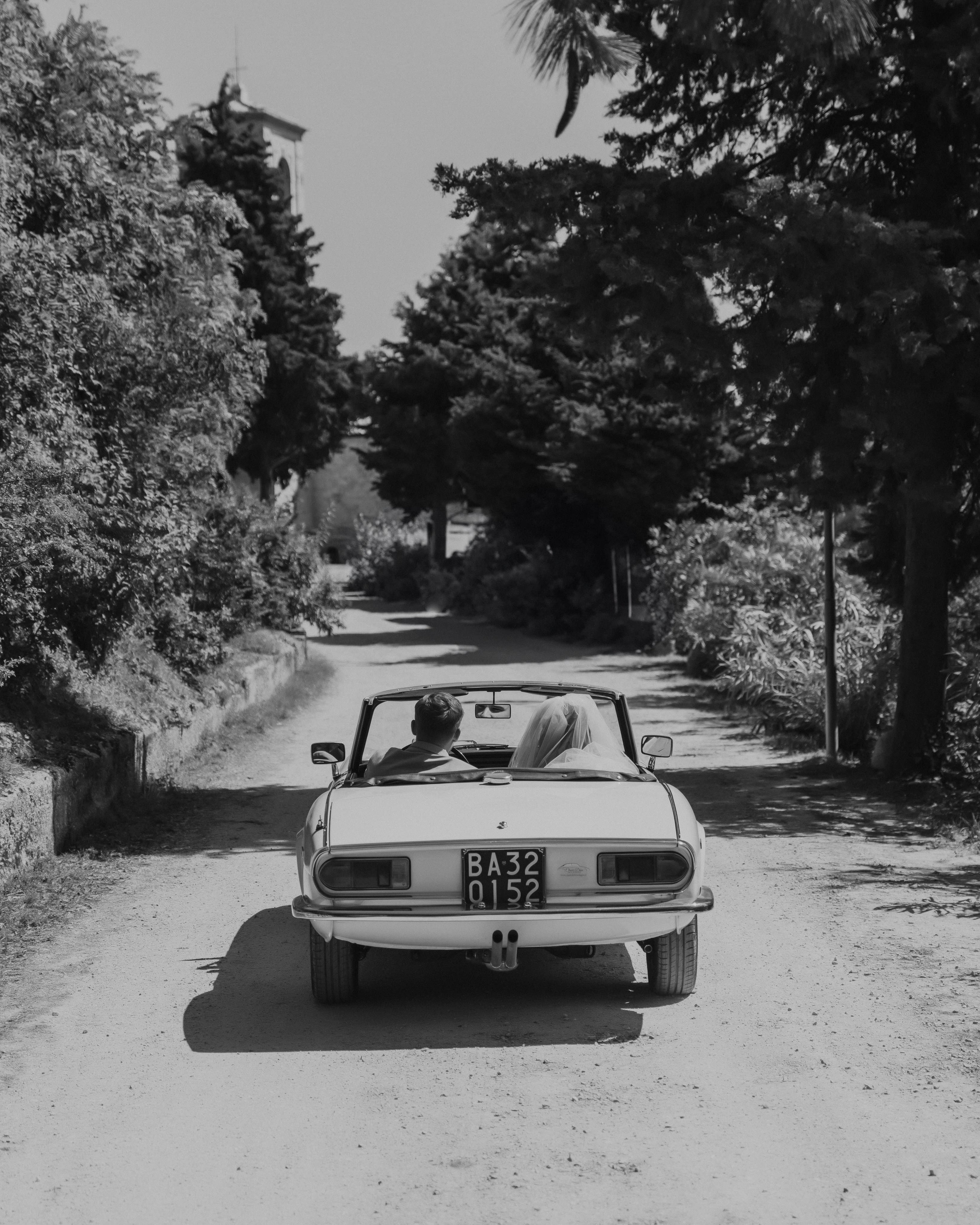 Noivos em carro clássico descapotável a sair da cerimónia, numa estrada com árvores centenárias.