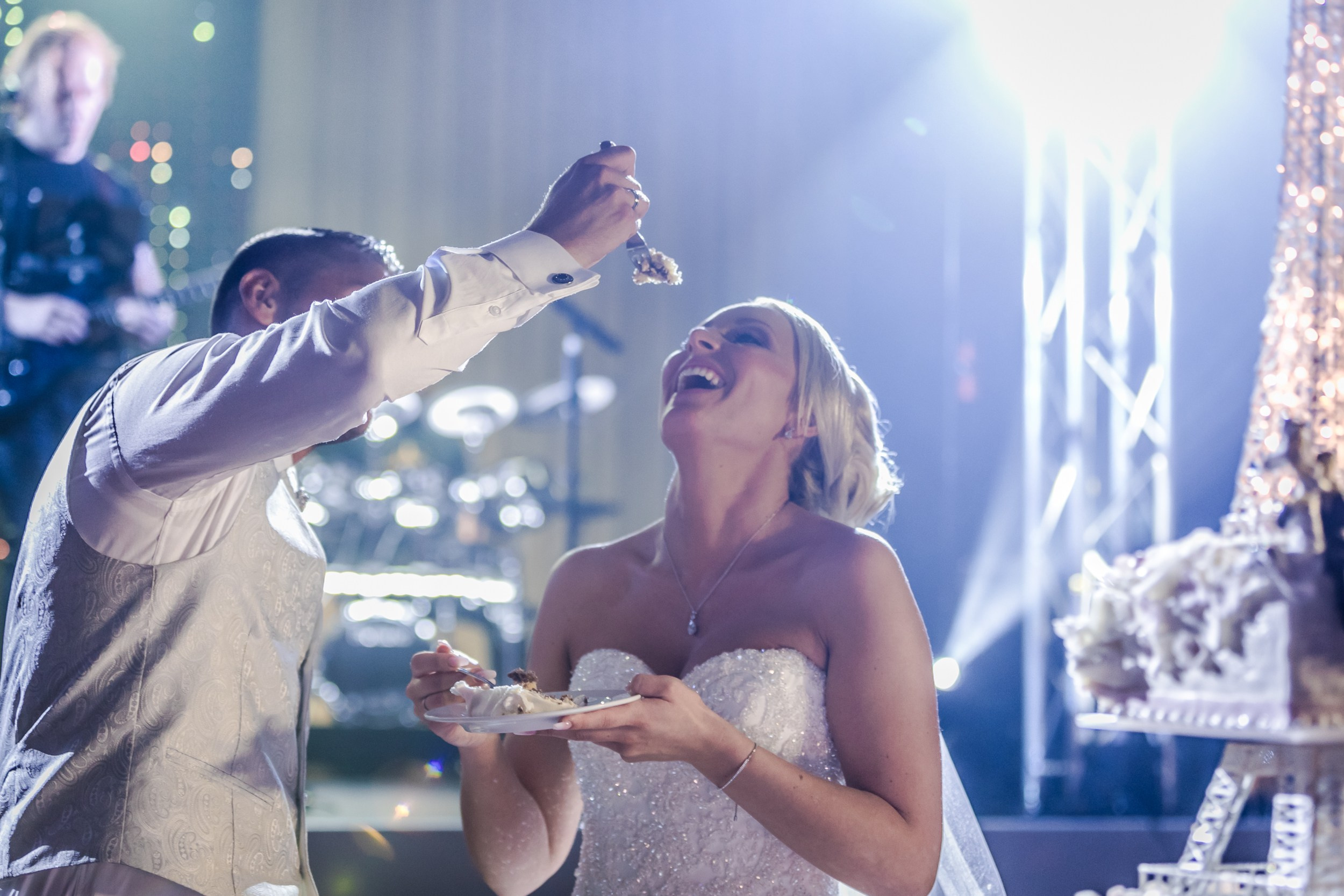 English:"Bride laughing joyfully as the groom playfully feeds her a bite of wedding cake during the reception. Bride in a sparkling strapless beaded wedding gown, groom in a light gray patterned vest and white shirt. Multi-tiered elegant wedding cake in the background with soft bokeh lights, capturing a candid and romantic moment at the wedding celebration."Русский:"Невеста радостно смеётся, пока жених шутливо кормит её кусочком свадебного торта на банкете. Невеста в сверкающем платье без бретелей с вышивкой бисером, жених в светло-сером узорчатом жилете и белой рубашке. На заднем плане многоярусный свадебный торт и мягкий боке от огней, трогательный и весёлый момент свадебного торжества."Deutsch:"Die Braut lacht herzlich, während der Bräutigam sie spielerisch mit einem Stück Hochzeitstorte füttert. Braut in einem funkelnden trägerlosen Brautkleid mit Perlenstickerei, Bräutigam in hellgrauer gemusterter Weste und weißem Hemd. Im Hintergrund die mehrstöckige elegante Hochzeitstorte und weiches Bokeh-Licht – ein fröhlicher und romantischer Moment auf der Hochzeitsfeier."
