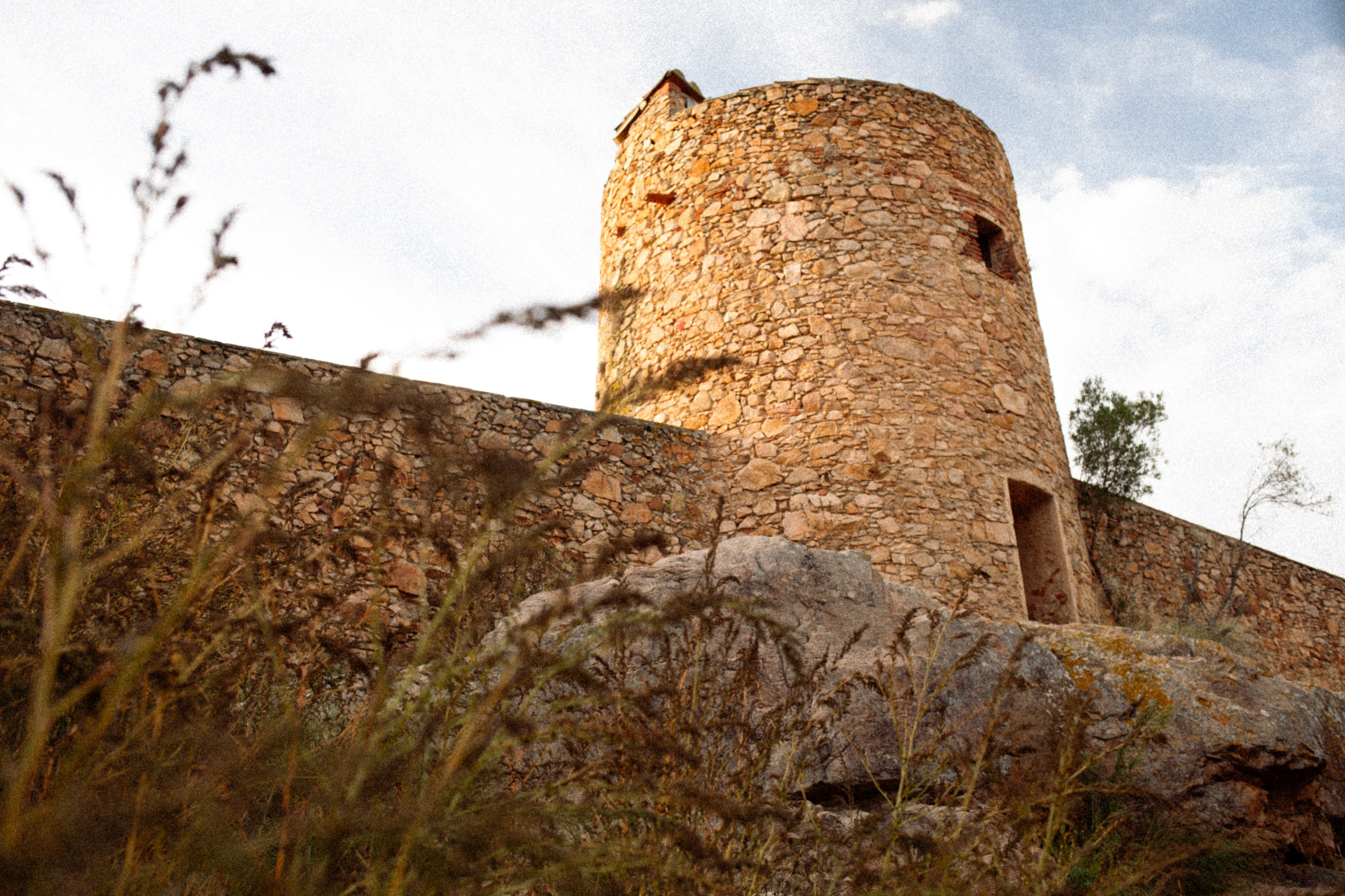 Historic fortress building in Can Duran bathed in sunset light, with dry grasses and wild plants in the foreground