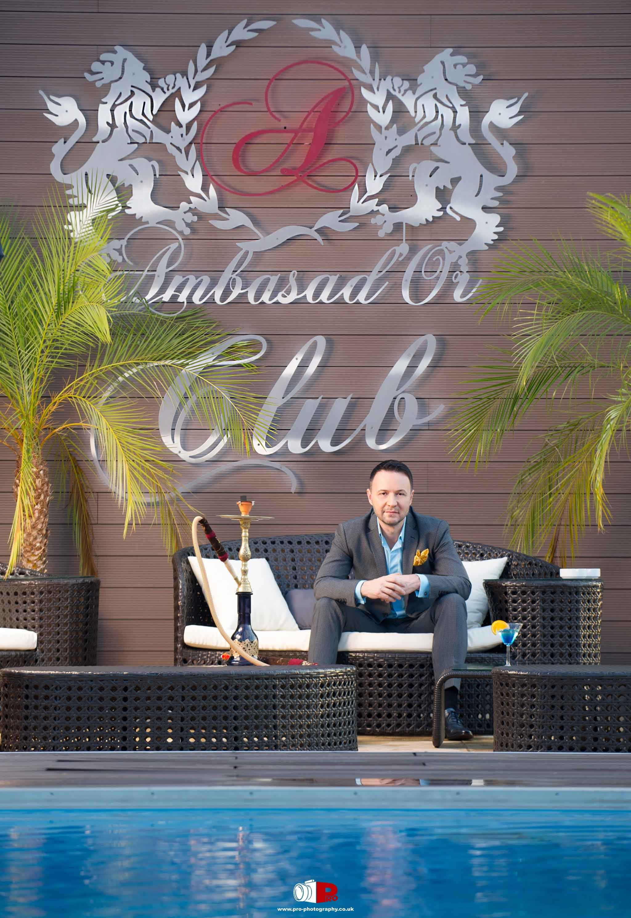 A man in a grey suit is relaxing on a stylish outdoor sofa by the pool at Ambasad'Or Club, which has palm trees and a hookah on display.