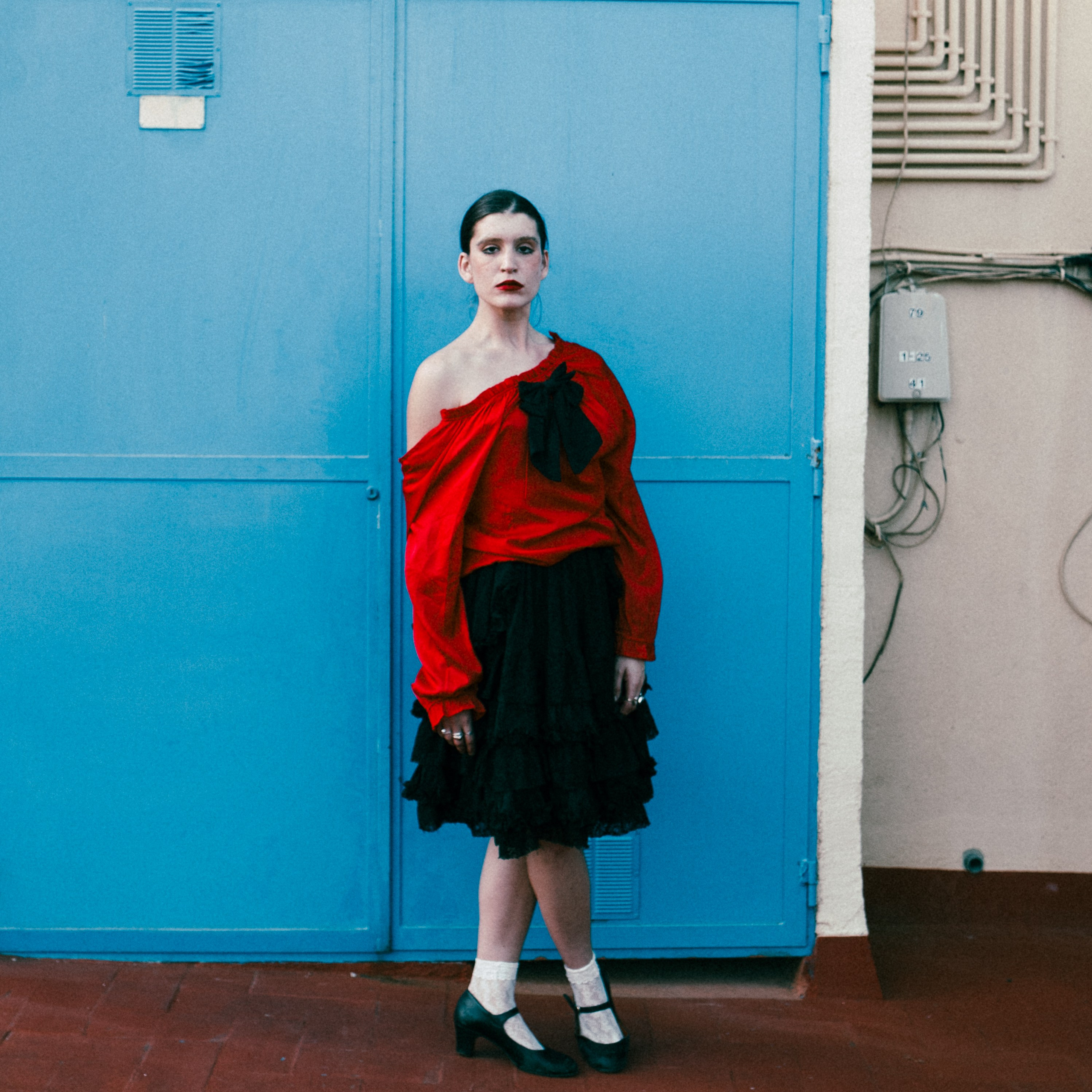 Young woman in red and black vintage outfit, standing against a blue wall