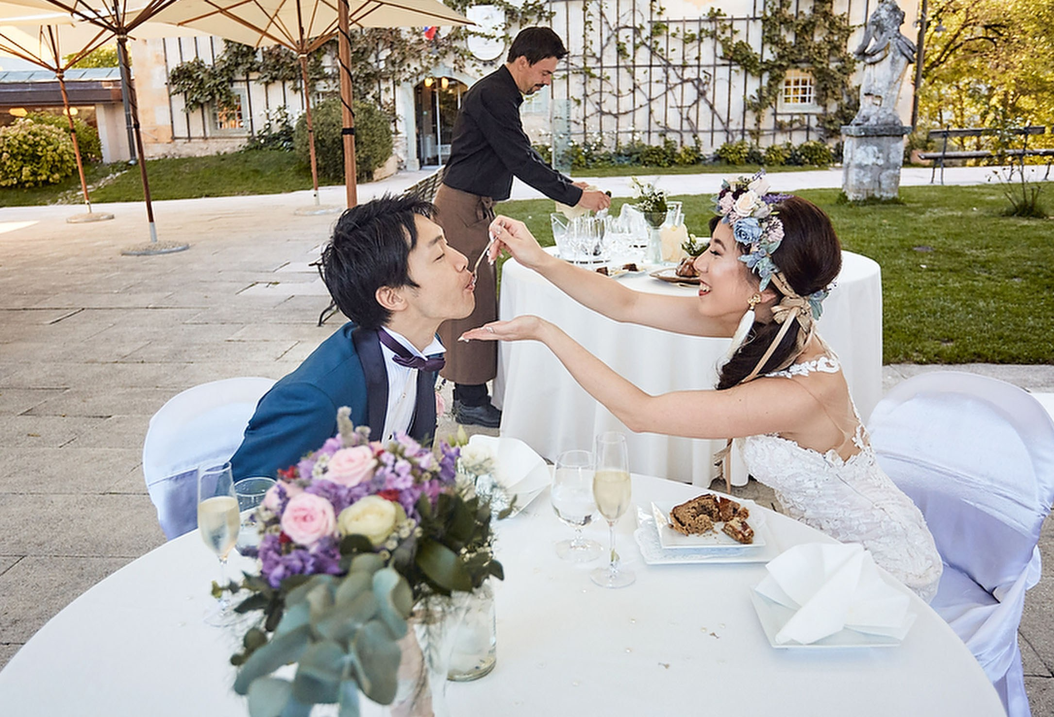 A smiling bride, wearing a floral headpiece feeds a blue-suited groom wedding cake in an outdoor setting.