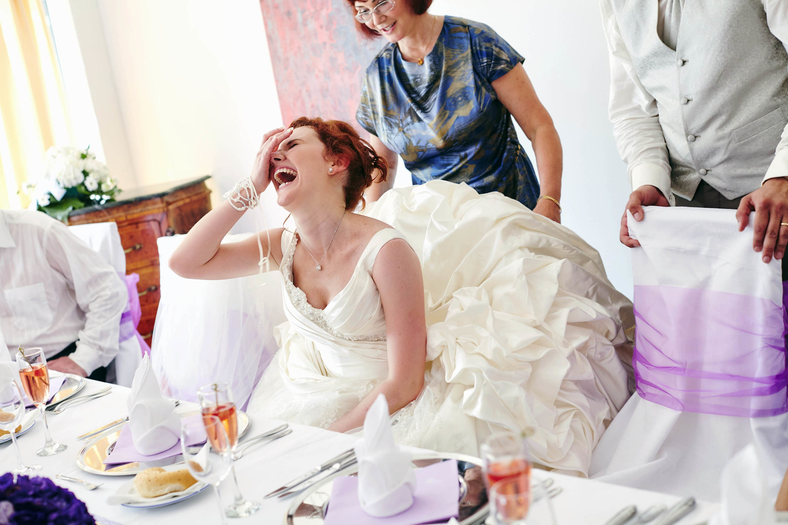 Red-haired bride laughing while mother helps with wedding dress at Castle Hluboka wedding dinner.