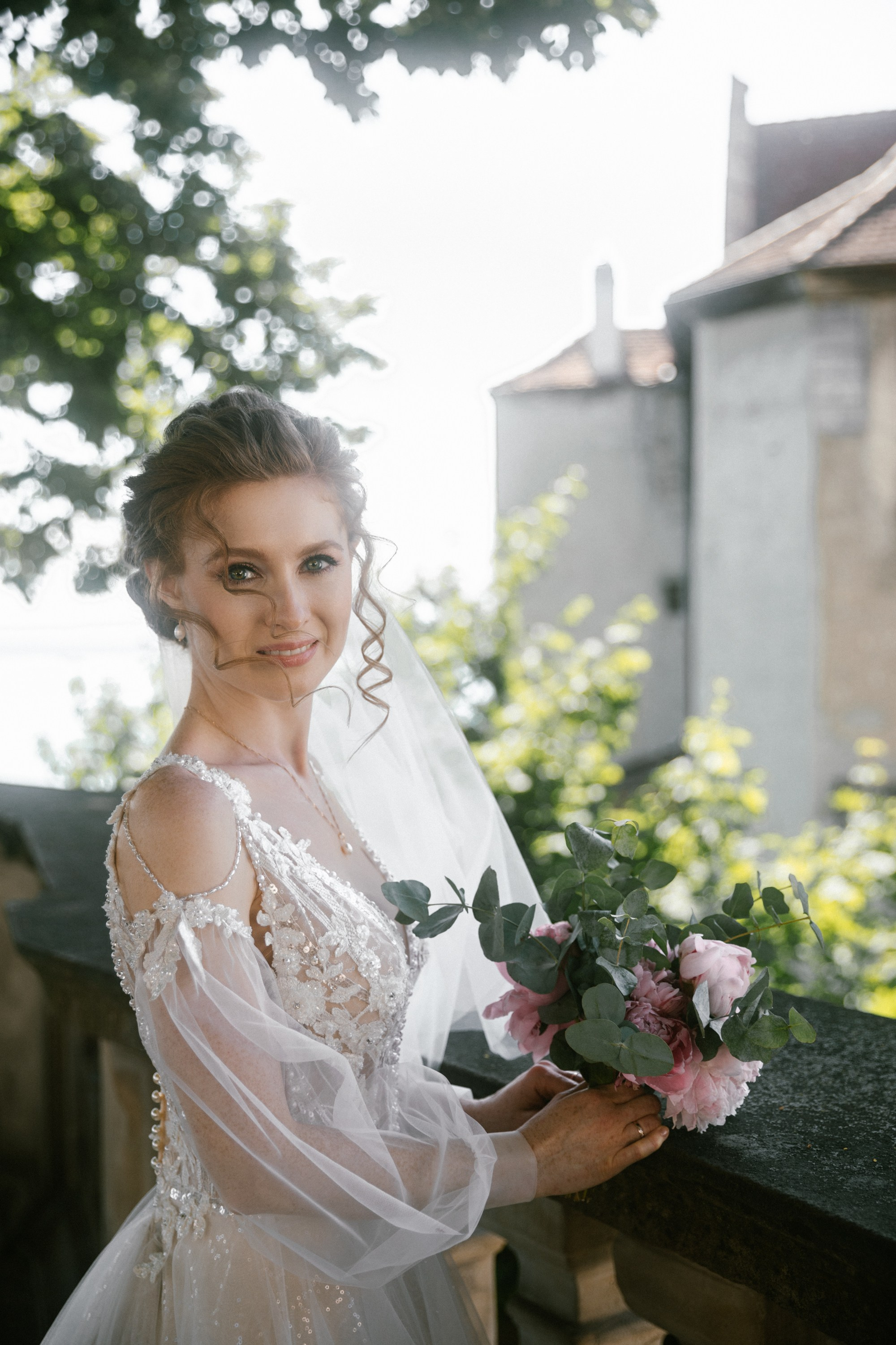 Bride standing alone on Schloss Meersburg balcony holding bouquet in sunlight