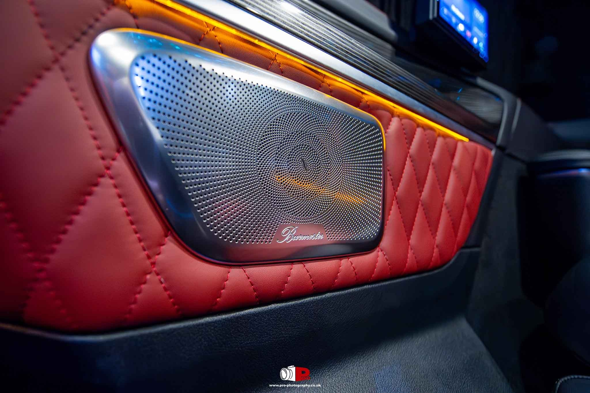Close-up of a Burmester speaker integrated into a luxurious red quilted car door panel with ambient lighting.