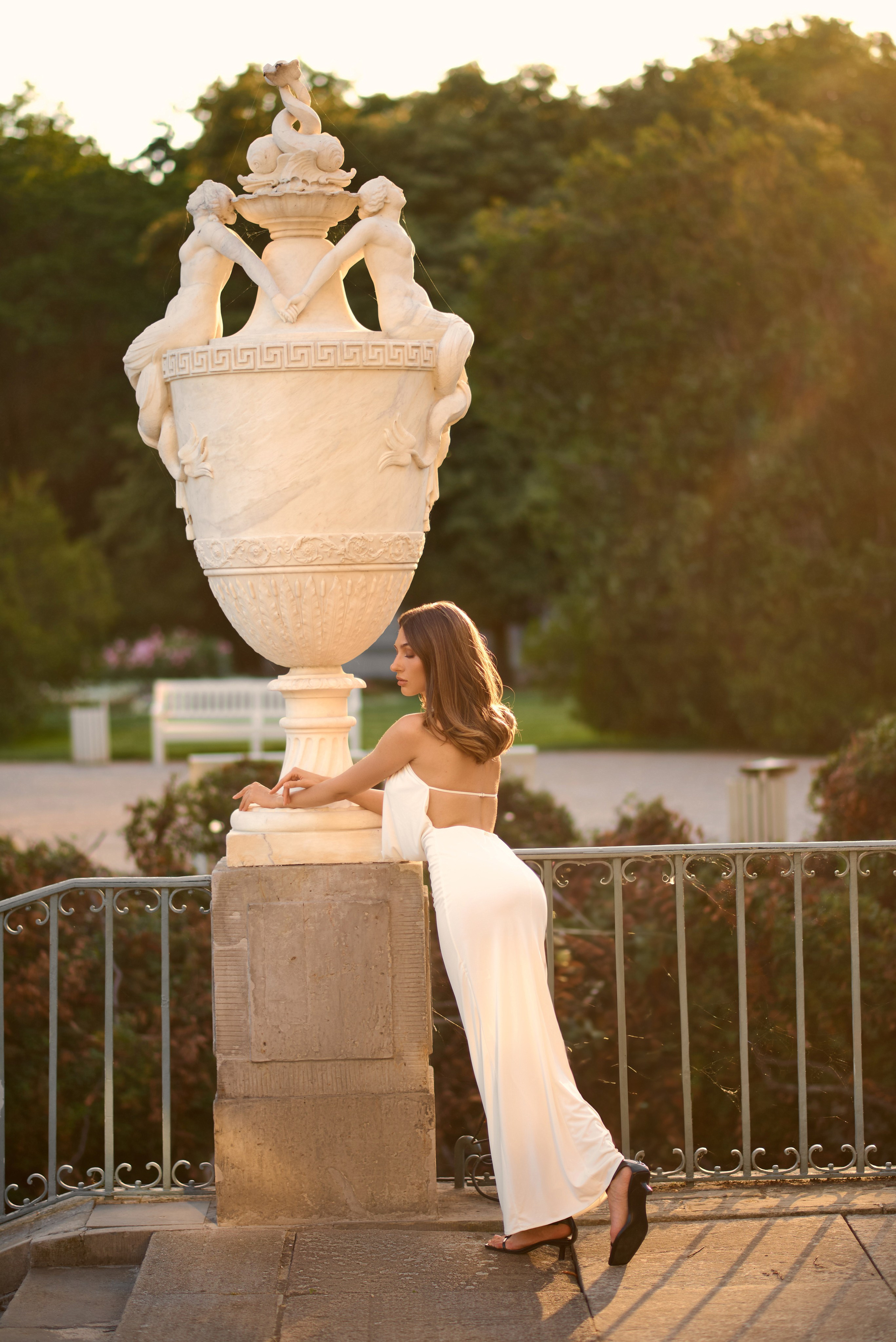 Жінка в облягаючій білій сукні з відкритою спиною нахиляється на огорожу, позуючи на тлі старовинної мармурової вази, увінчаної фігурами русалок.