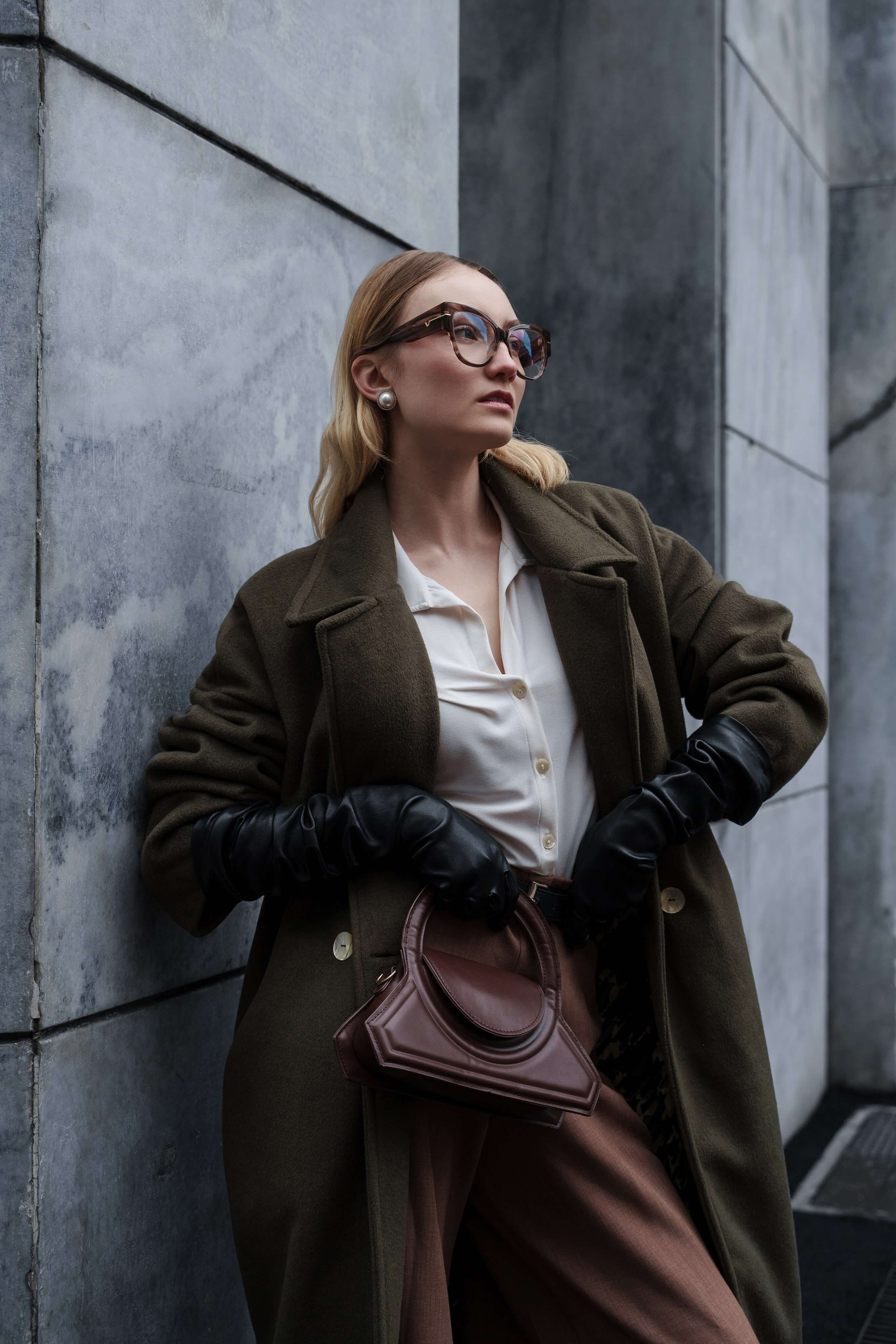 Fashion model with tortoiseshell glasses and pearl earrings posing in an olive coat and brown trousers