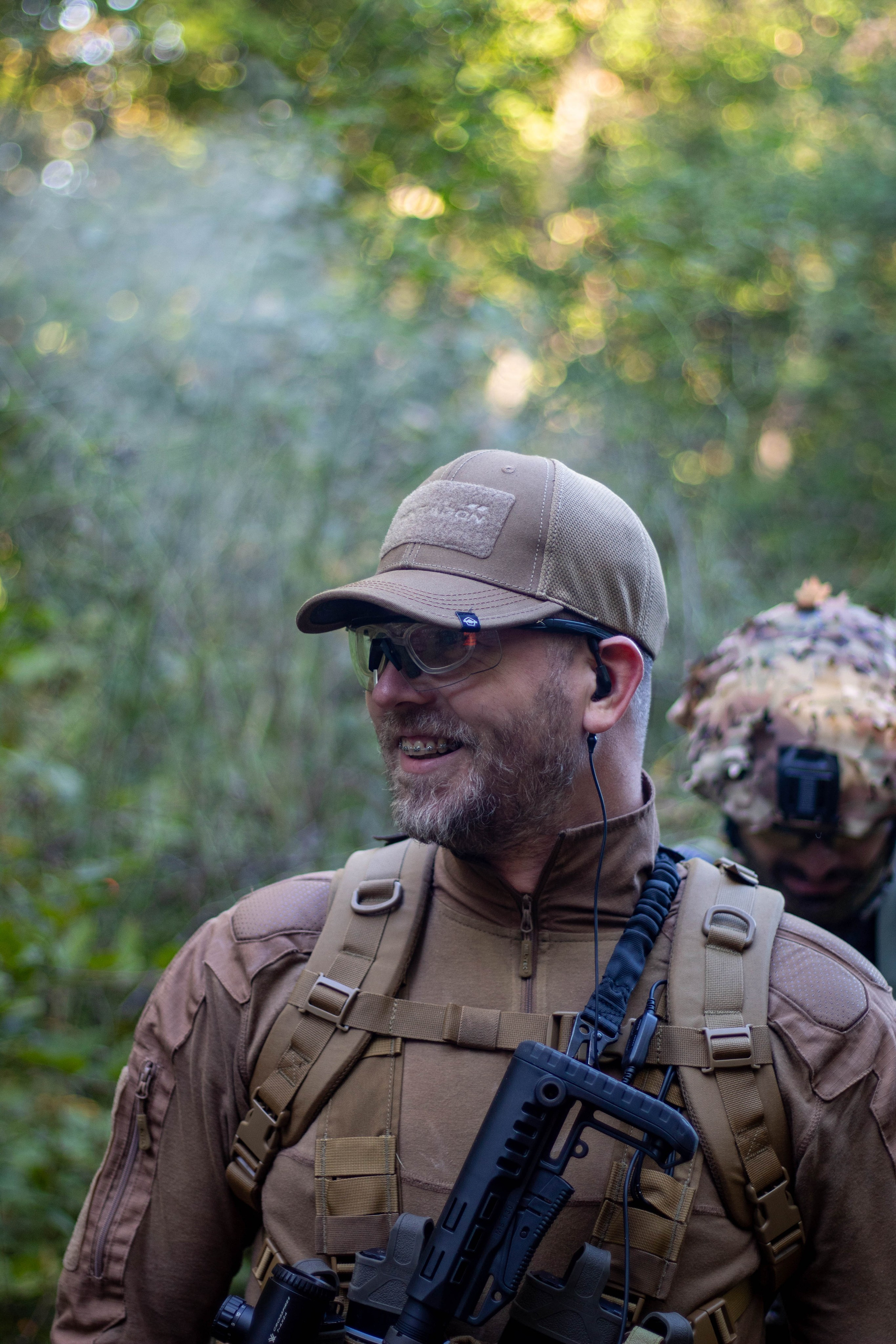 Portrait of a man in tactical military-style gear holding a rifle in a forested area.