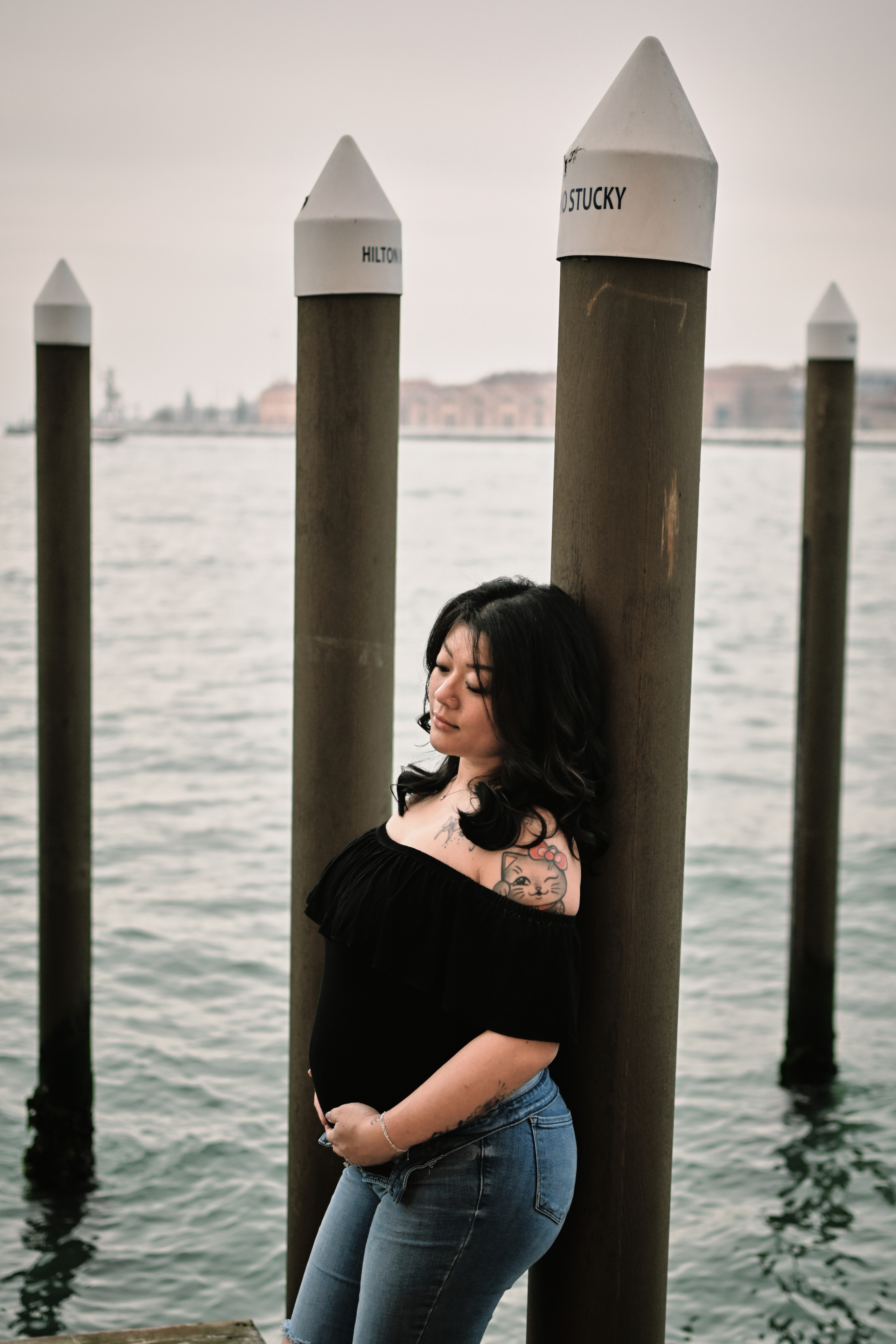 A pregnant young woman stands on a bridge in Venice, wearing a pair of comfortable jeans and a flowing white blouse. Her hair is pulled back in a loose bun, and she gazes out at the bustling canal below. The sunlight glimmers on the water, casting a warm glow over the scene. In the background, ancient buildings and colorful boats lend a sense of history and romance to the setting. The woman's pose is relaxed yet confident, conveying her excitement and anticipation for the new life growing inside her.
