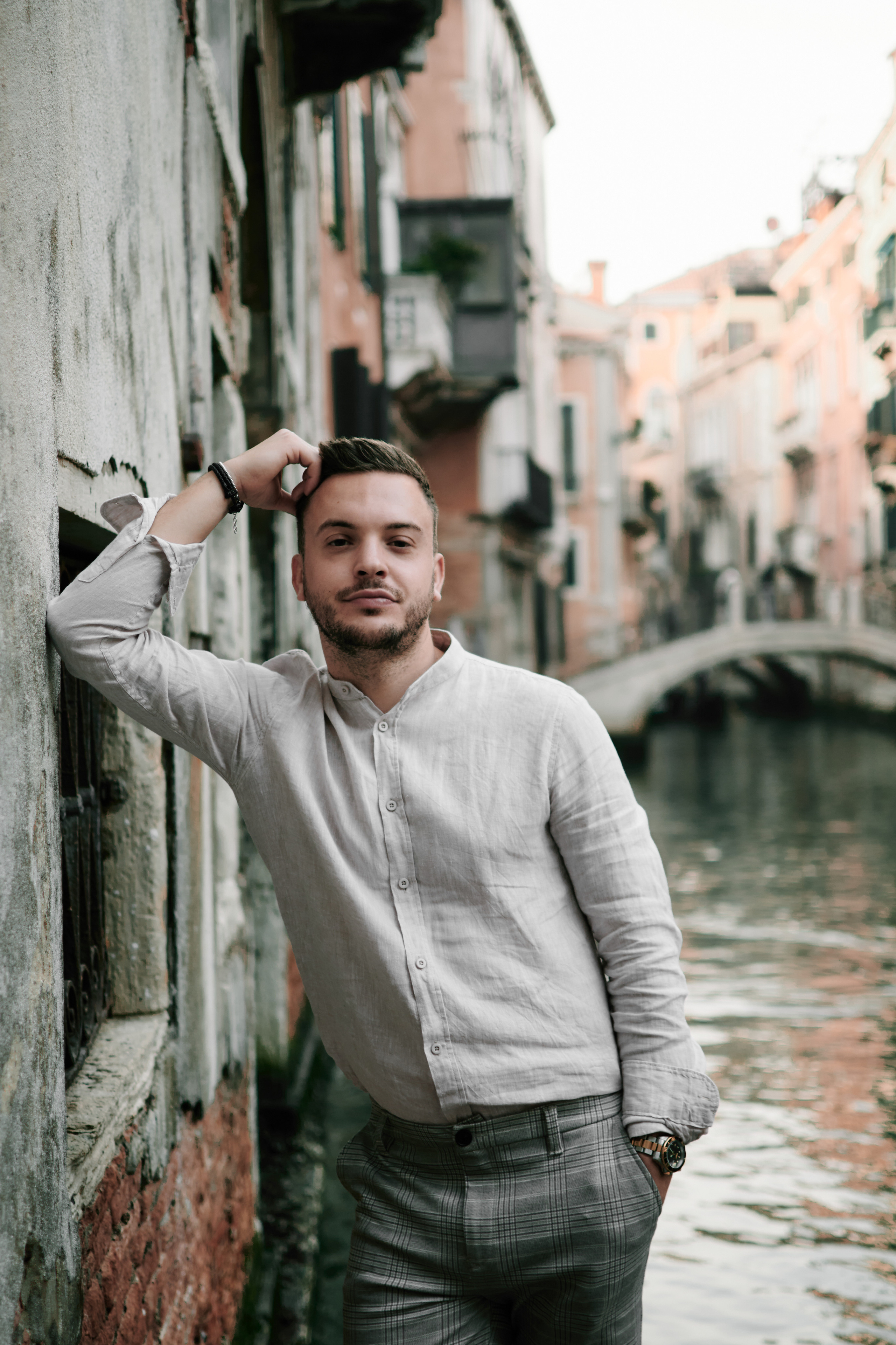 A young man stands on a narrow street in Venice, Italy. The sun casts a warm glow on the pastel-colored buildings, creating a picturesque backdrop. The man looks out into the distance, his expression contemplative and serene. The narrow street is lined with quaint shops and cafes, adding to the charm of the scene. The man's outfit is stylish and modern, yet it blends seamlessly with the timeless beauty of Venice. The photo captures a moment of quiet reflection amidst the bustling city, evoking a sense of peacefulness and introspection
