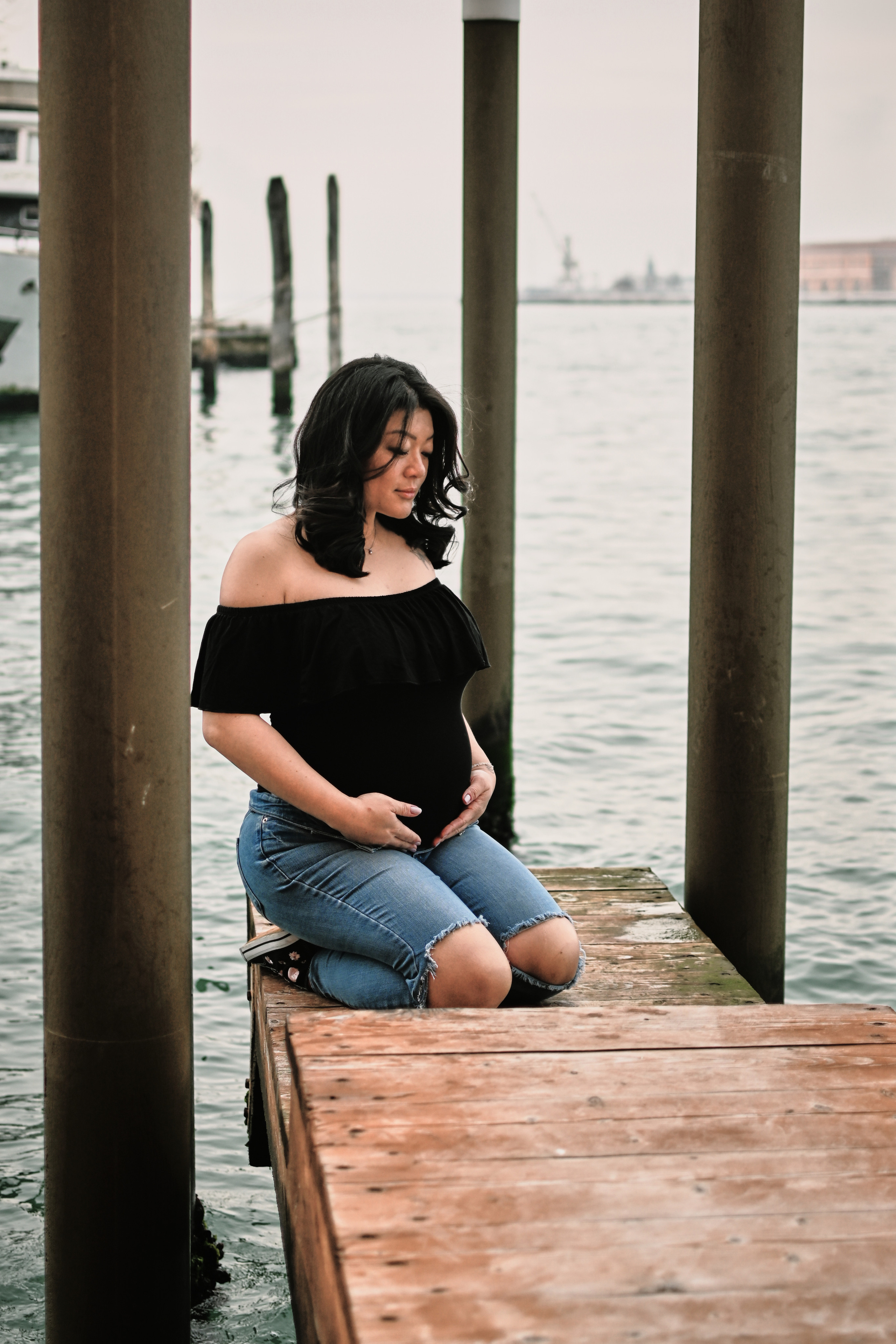 A pregnant young woman stands on a bridge in Venice, her hands cradling her belly. The water of the canal below reflects the warm light of the setting sun, casting a golden glow over the entire scene. Despite the bustling crowds around her, the woman seems lost in thought, her expression a mix of anticipation and nervousness. Her gaze is fixed on the horizon, as if awaiting the arrival of someone very special