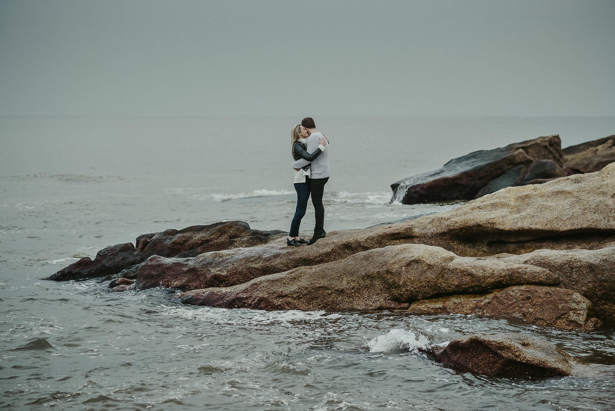 Мужчина обнимает любимую стоя на камнях у прибоя