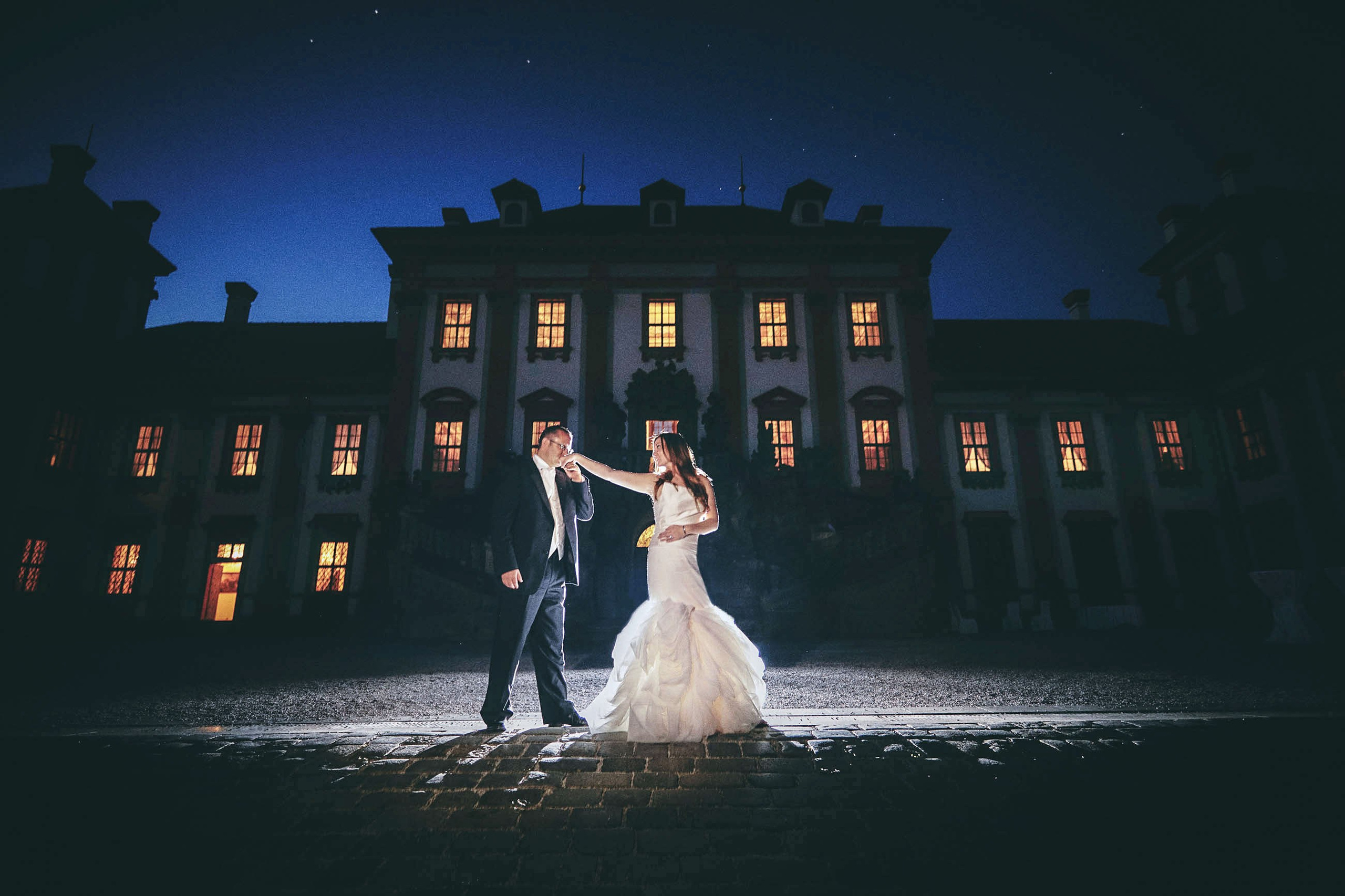 Dallas groom kissing bride's hand under the night-lit facade of Troja Chateau, illuminated by off-camera flash.