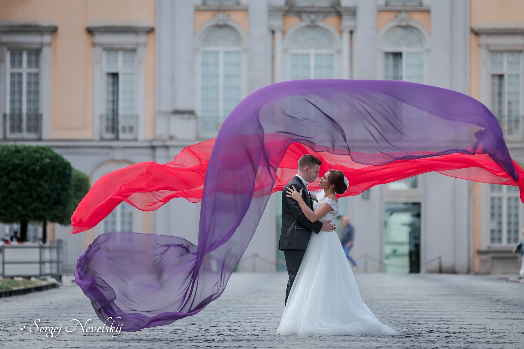 English:"Romantic newlyweds embracing under a flowing red-to-purple veil billowing dramatically in the wind. Bride in an elegant white lace wedding dress with off-the-shoulder sleeves, groom in a classic black tuxedo. Historic European palace with arched windows in the background, creating a fairytale-like wedding portrait. Photo by © Sergej Nevelskij"Русский:"Молодожёны нежно обнимаются под развевающейся на ветру яркой красно-фиолетовой вуалью. Невеста в элегантном белом свадебном платье с открытыми плечами и кружевом, жених в классическом чёрном смокинге. На заднем плане исторический дворец с арочными окнами — сказочный свадебный портрет. Фото © Sergej Nevelskij"Deutsch:"Verliebtes Brautpaar umarmt sich unter einem dramatisch im Wind wehenden rot-violetten Schleier. Braut in einem eleganten weißen Brautkleid mit Carmen-Ausschnitt und Spitze, Bräutigam im klassischen schwarzen Smoking. Im Hintergrund ein historisches Schloss mit Rundbogenfenstern – ein märchenhaftes Hochzeitsporträt. Foto © Sergej Nevelskij"