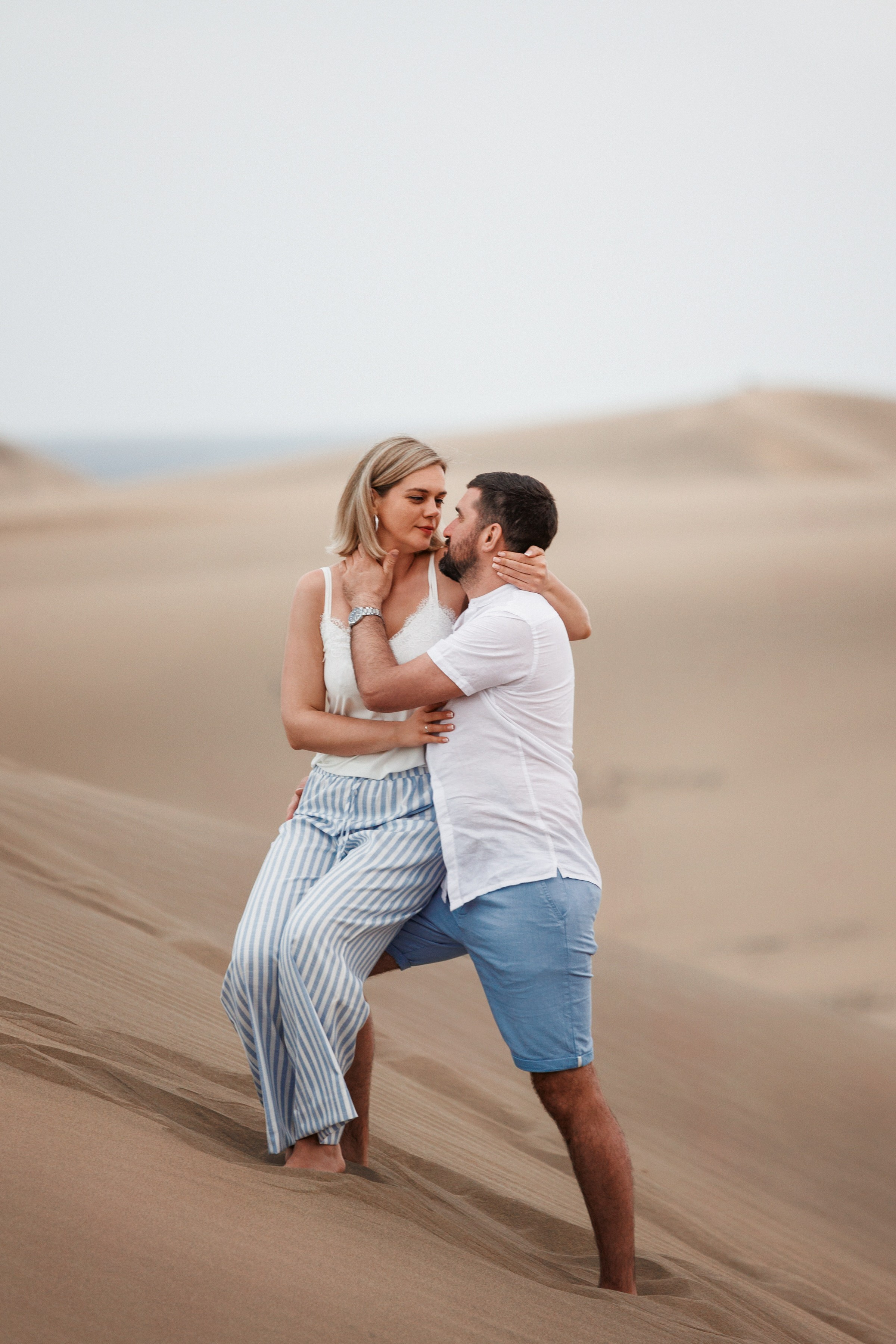 Love Story Photographer - A couple embracing in a sandy desert, with the man kissing the woman's cheek. both are dressed in light, casual summer clothing.