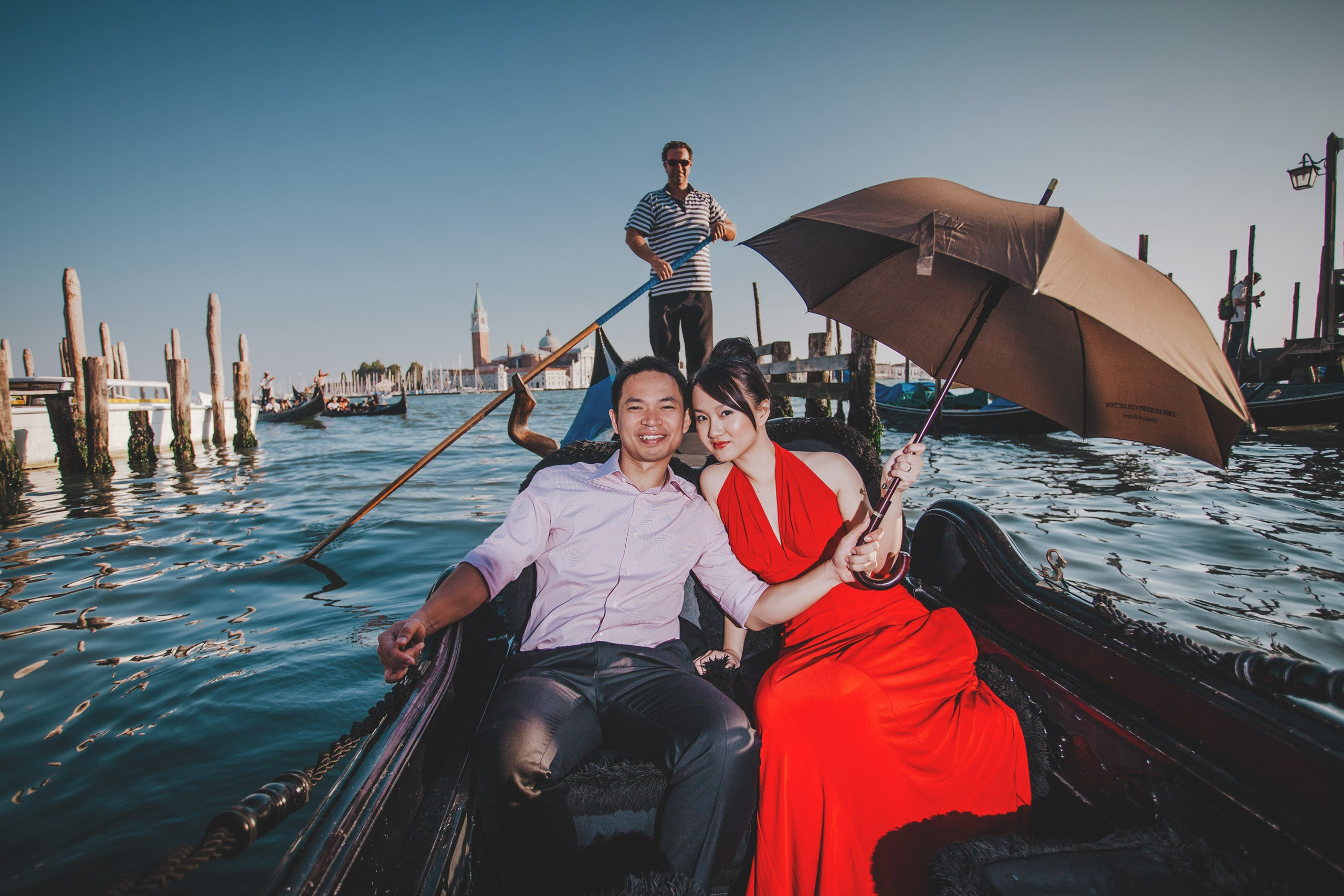 A smiling Thai woman wearing a sexy red evening dress and her fiancee holding an umbrella embark on a gondola ride in the canals of Venice.