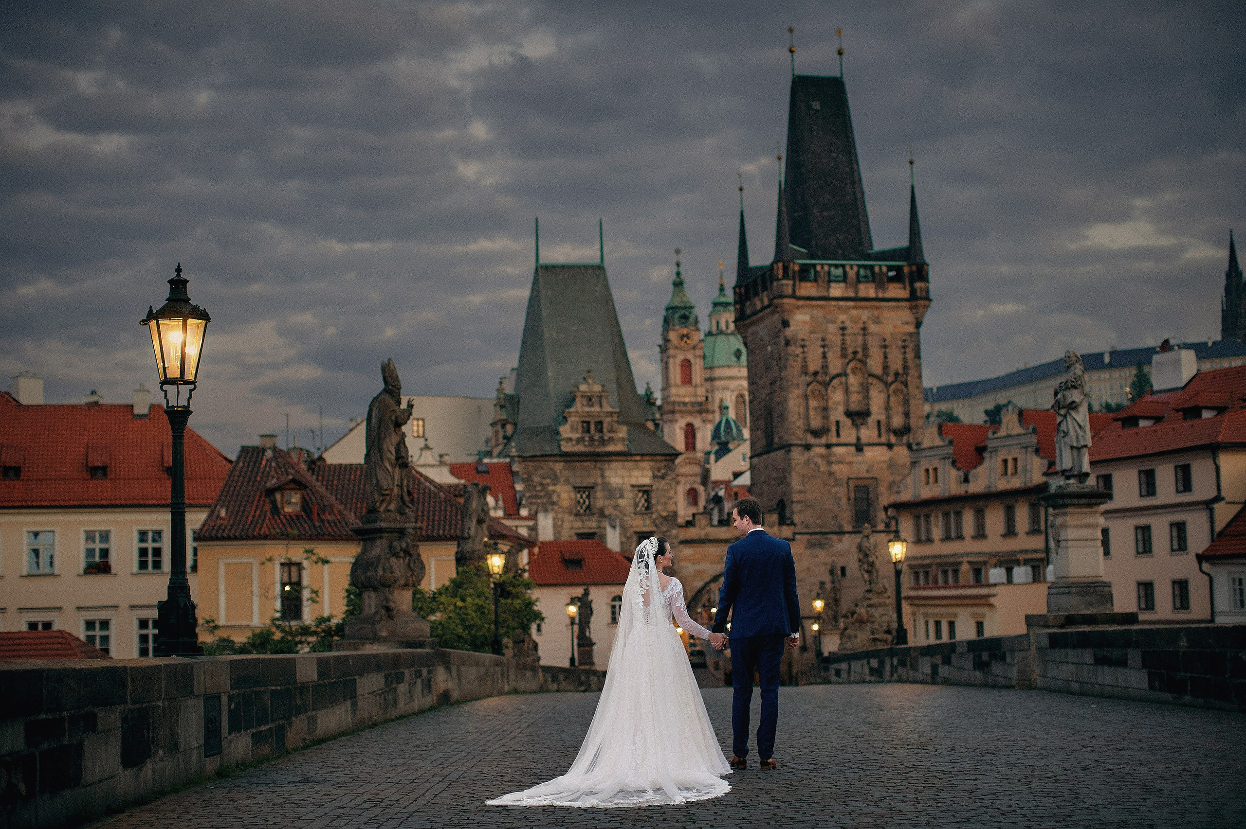 A bride and groom walk hand in hand across a deserted Charles Bridge illuminated by its historic gas lamps during their destination wedding in Prague.Newlyweds stand underneath a designer floral arch created in their honor as the twinkling lights of Prague is seen below them.Adéla & Matthias