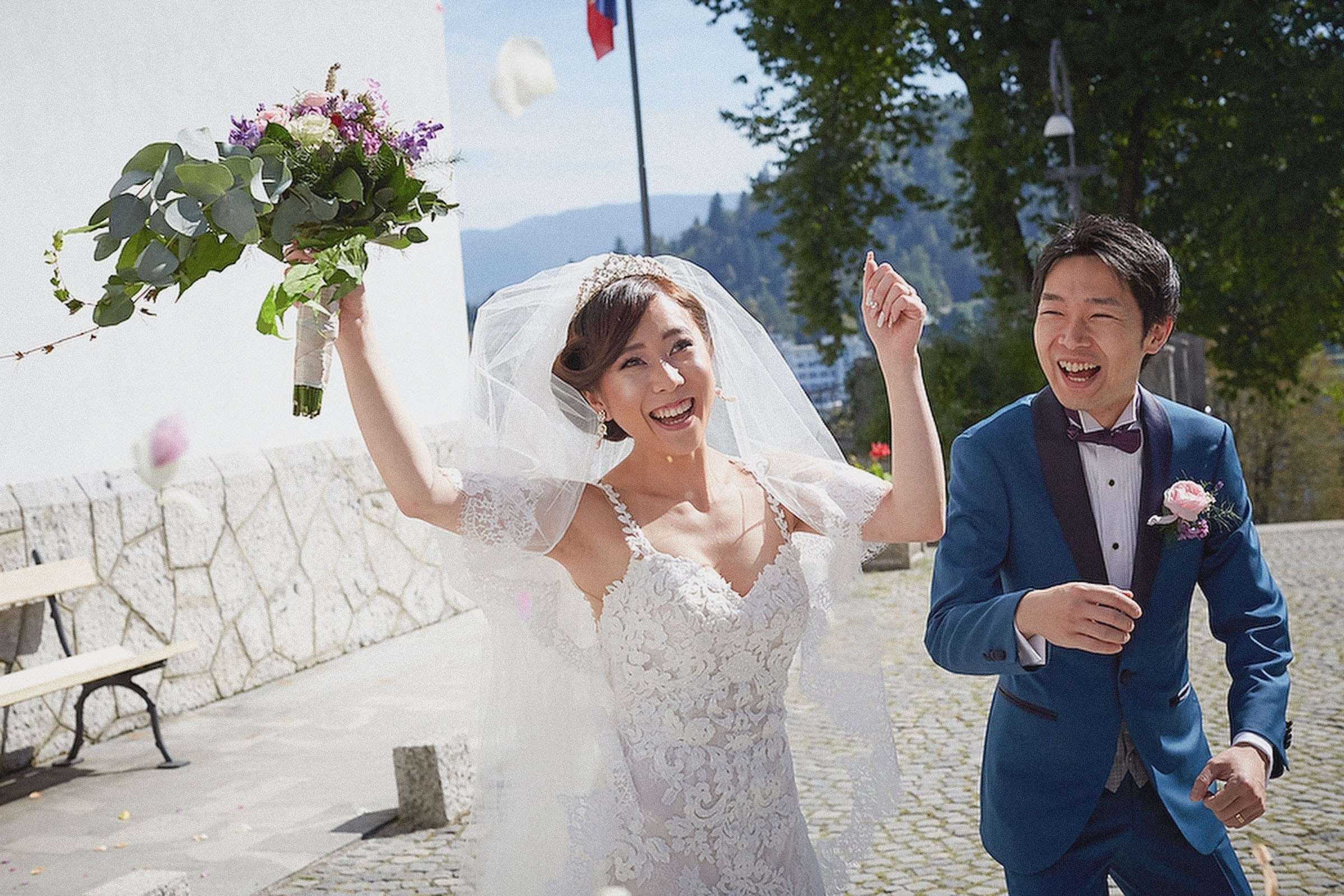 A young Japanese bride holds her arms in the air in a jubilant pose after exiting a church with her smiling groom.