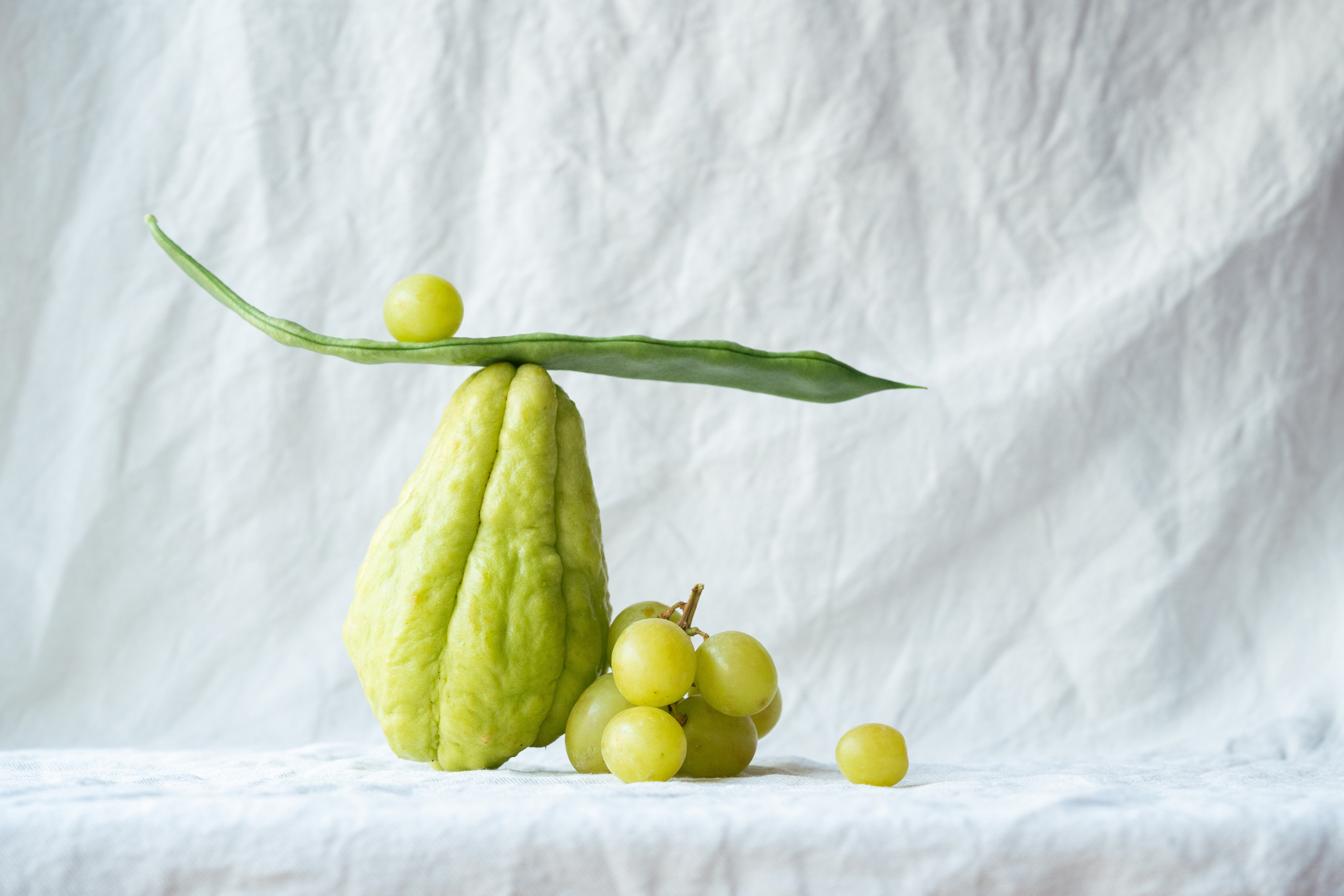 Ainda vida equilibrada de Chayote, feijão verde e uvas. Arranjo artístico de chayote fresco, feijão verde e uvas equilibradas em um fundo de tecido texturizado branco.