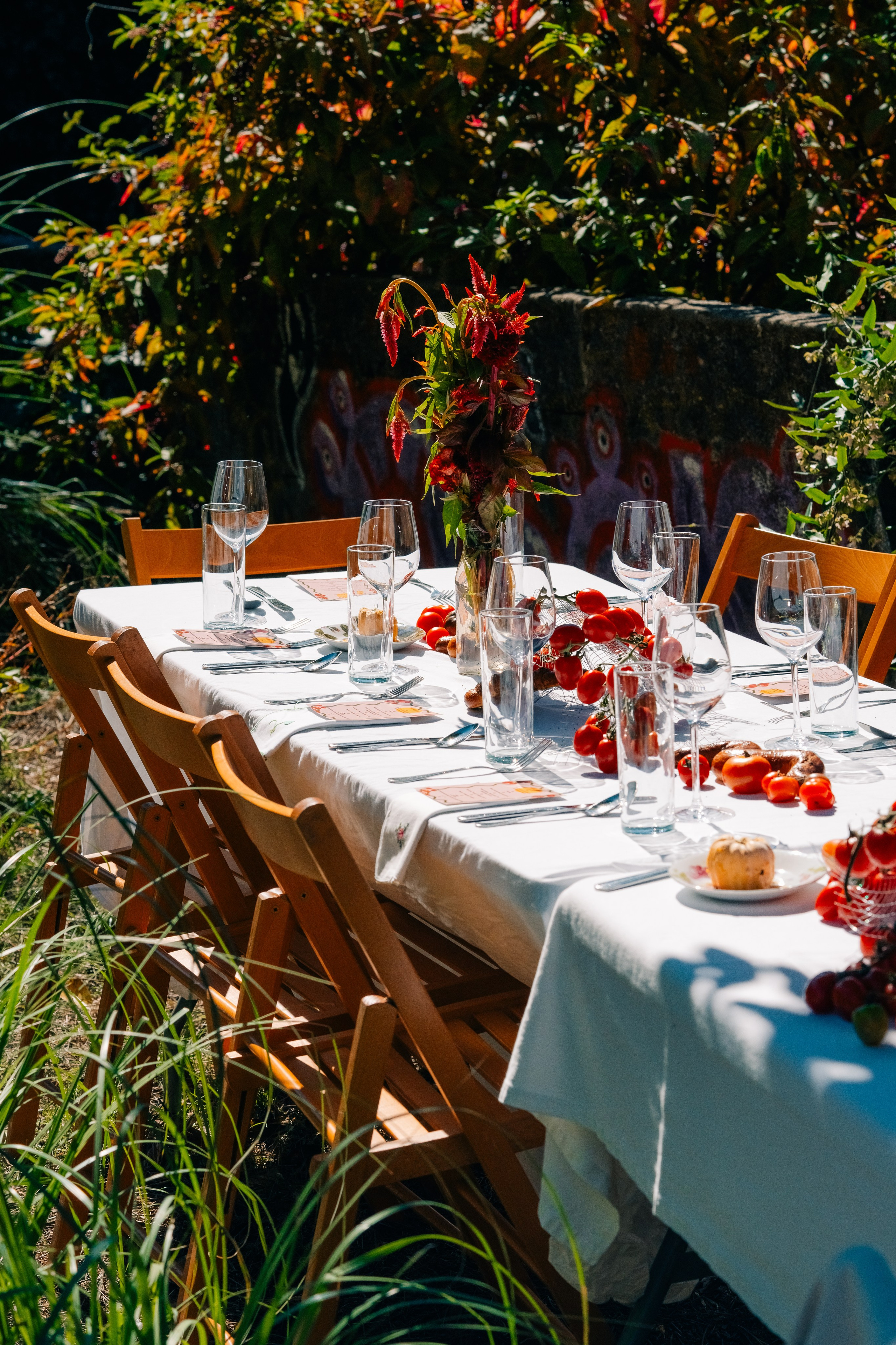 Configuração elegante de mesa de exterior com peça central floral num jardim. Mesa de jantar ao ar livre lindamente decorada com peça central floral, copos e cadeiras de madeira situada num jardim.
