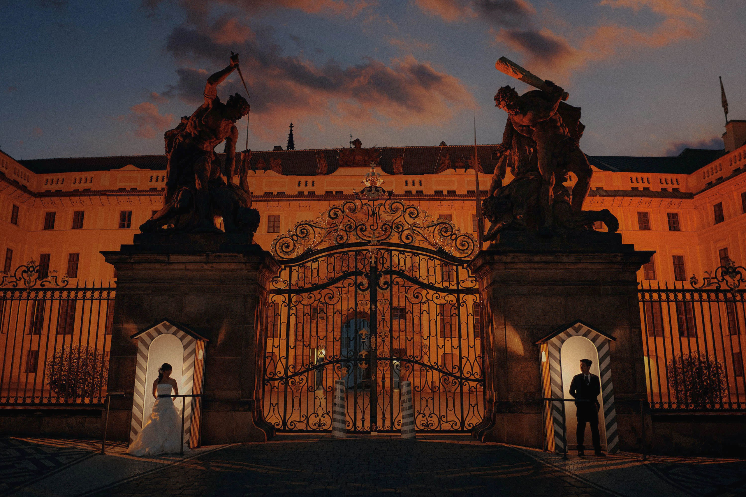 Playful bride and groom posing as castle guards in the guard shacks at Prague Castle, with a brilliantly flaring early morning sky overhead.