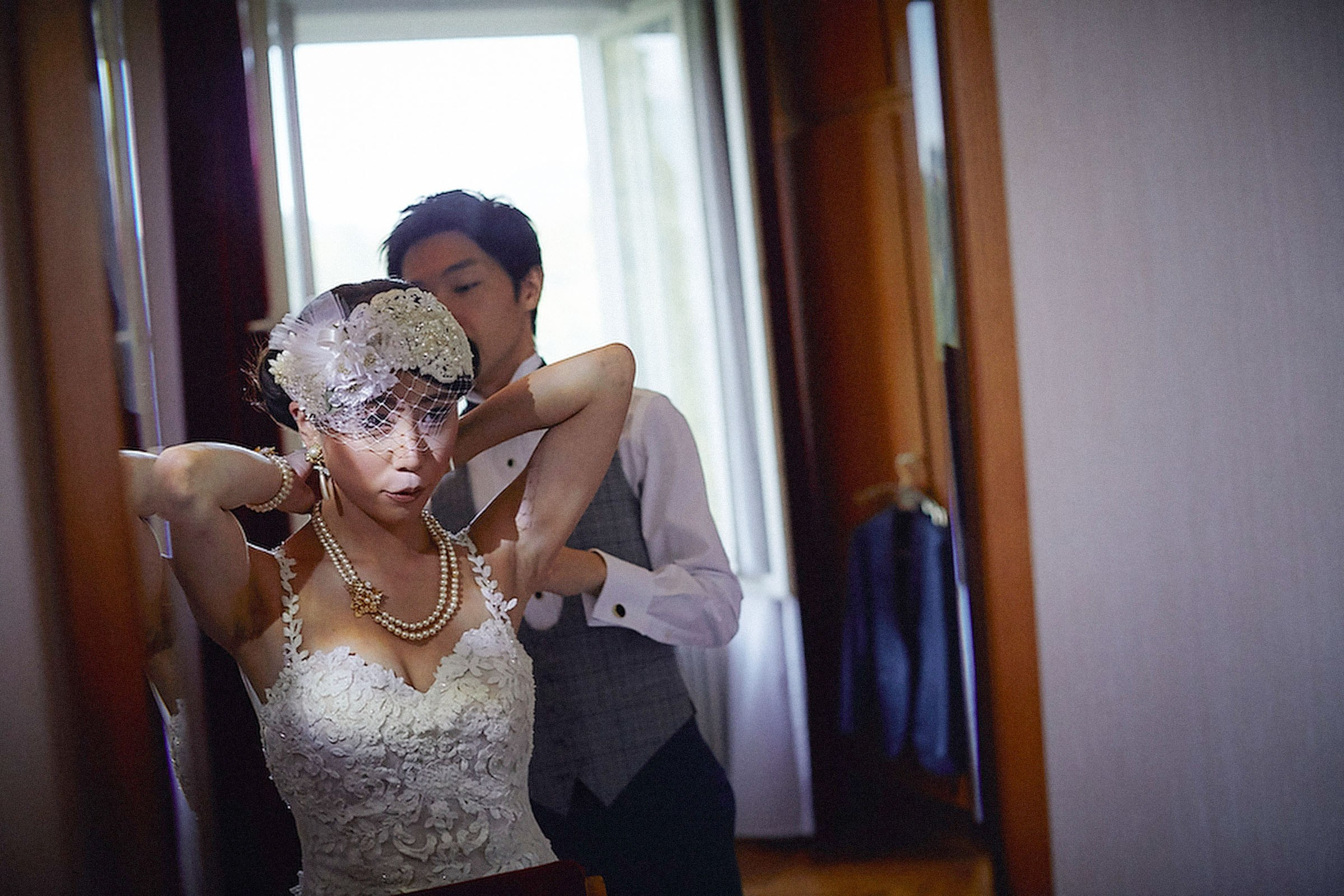 A groom assists the bride in putting on her jewelry in their suite.