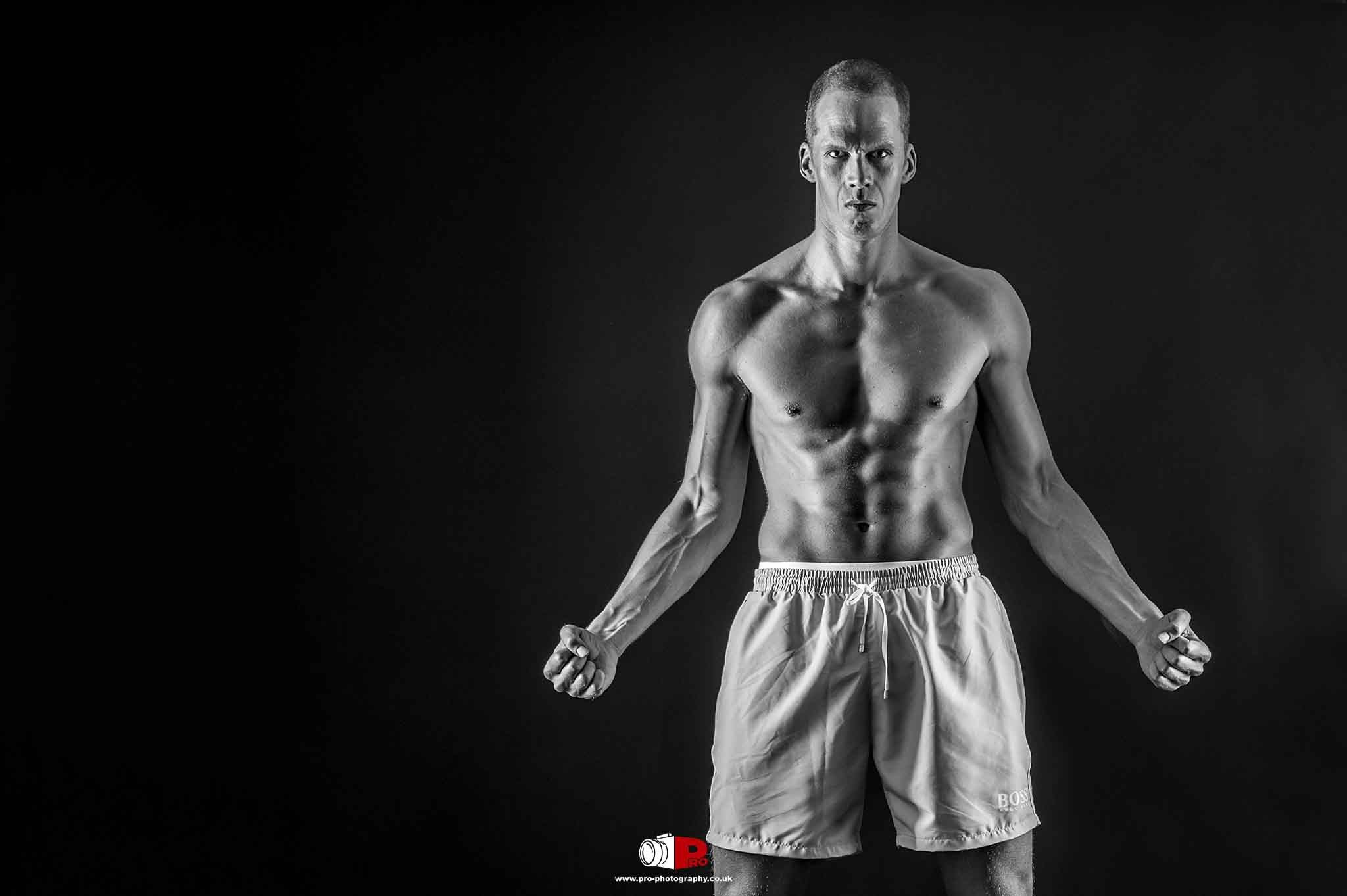 Muscular male fitness model in black and white posing shirtless under dramatic lighting.