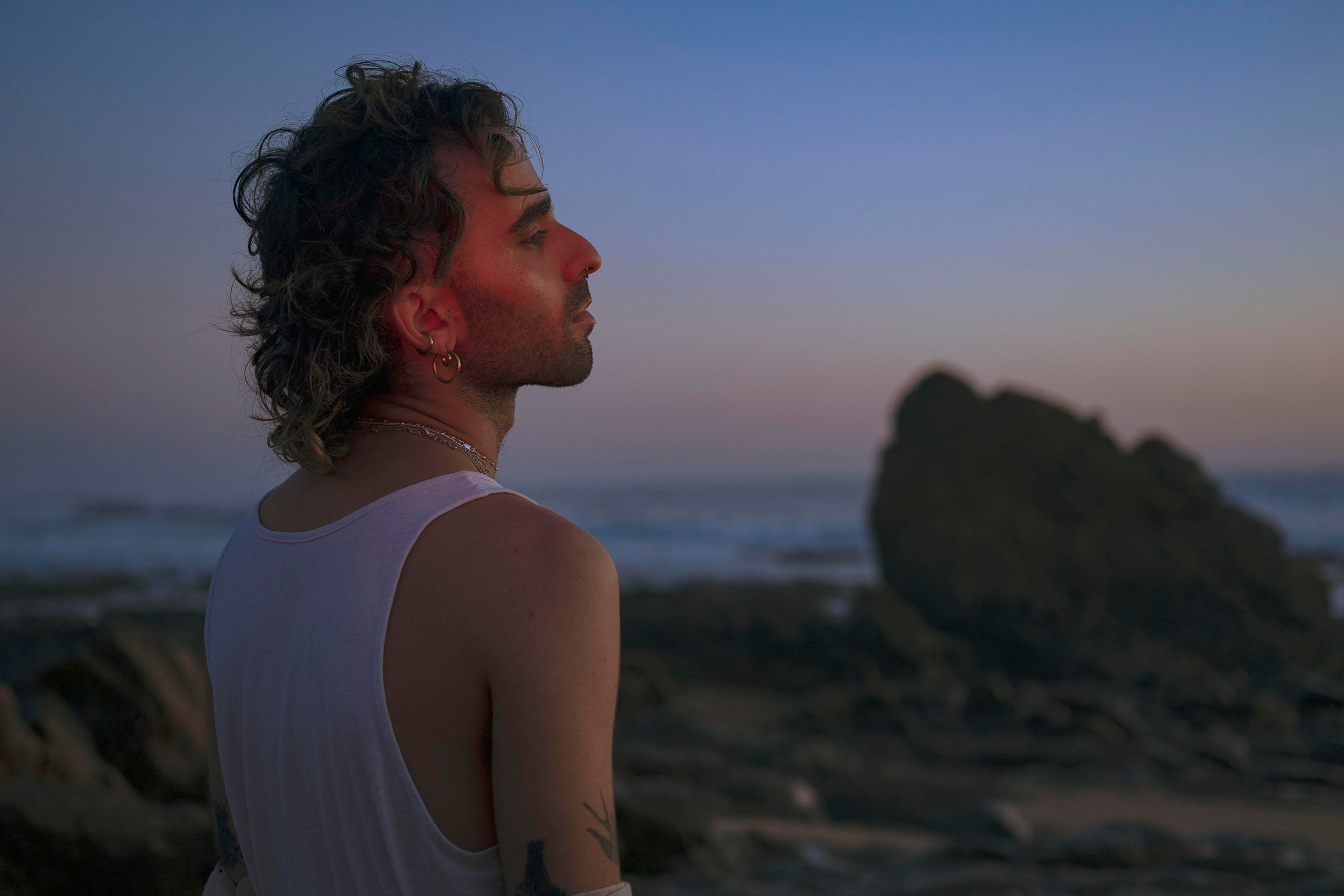 LGBTQ+, LGBT portrait of a beautiful person with makeup, a white tank top, and curly hair. Sunset on a beach, colored light.