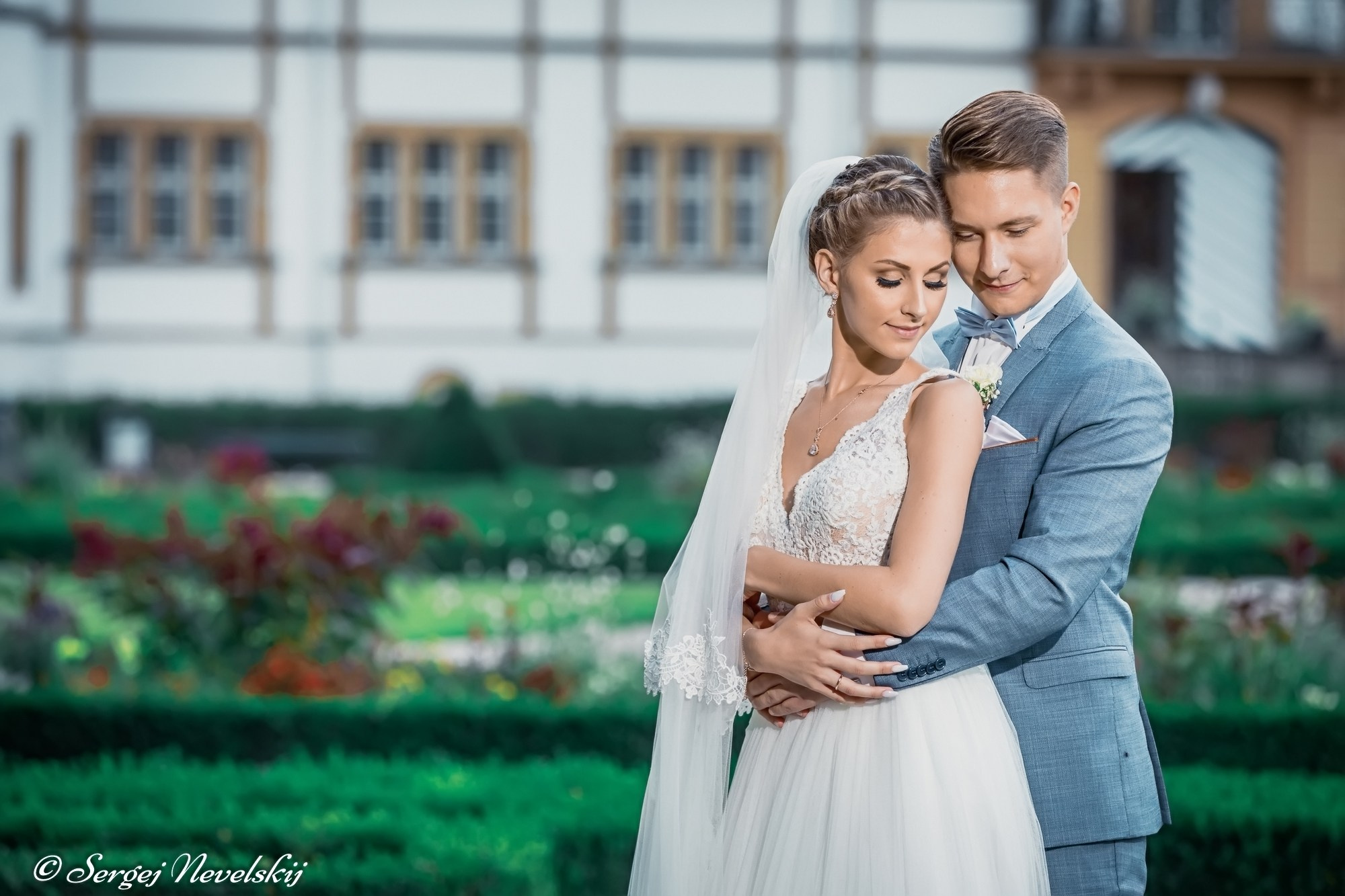 English:Tender moment of newlyweds embracing in front of a historic half-timbered palace. Bride in an elegant white lace wedding dress with deep V-neckline and long veil, groom in a light blue suit with matching bow tie and white rose boutonniere. Soft natural light, lush garden backdrop, capturing pure love and serenity. Photo by © Sergej NevelskijРусский:Нежный момент: молодожёны обнимаются на фоне старинного фахверкового дворца. Невеста в изящном белом кружевном свадебном платье с глубоким вырезом и длинной фатой, жених в голубом костюме с бабочкой и бутоньеркой из белой розы. Мягкий свет, цветущий сад — чистая любовь и умиротворение. Фото © Sergej NevelskijDeutsch:Zärtlicher Augenblick: Brautpaar umarmt sich vor einem historischen Fachwerk-Schloss. Braut in einem eleganten weißen Spitzenbrautkleid mit tiefem V-Ausschnitt und langem Schleier, Bräutigam im hellblauen Anzug mit passender Fliege und weißer Rosen-Ansteckblume. Weiches Licht, blühender Garten – pure Liebe und Ruhe. Foto © Sergej Nevelskij