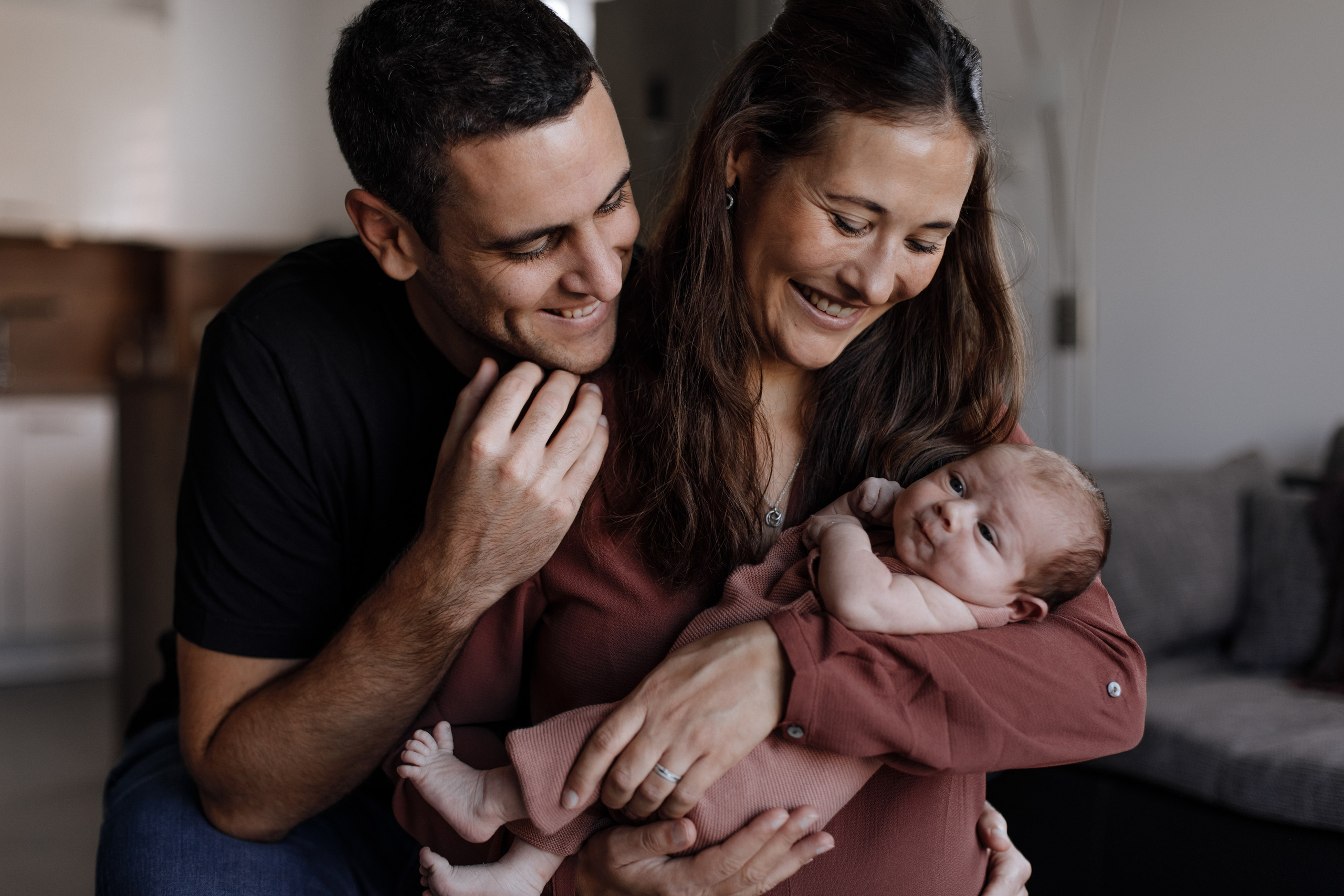 Mama hält Baby im Arm, während Papa sie liebevoll an den Schultern umarmt.